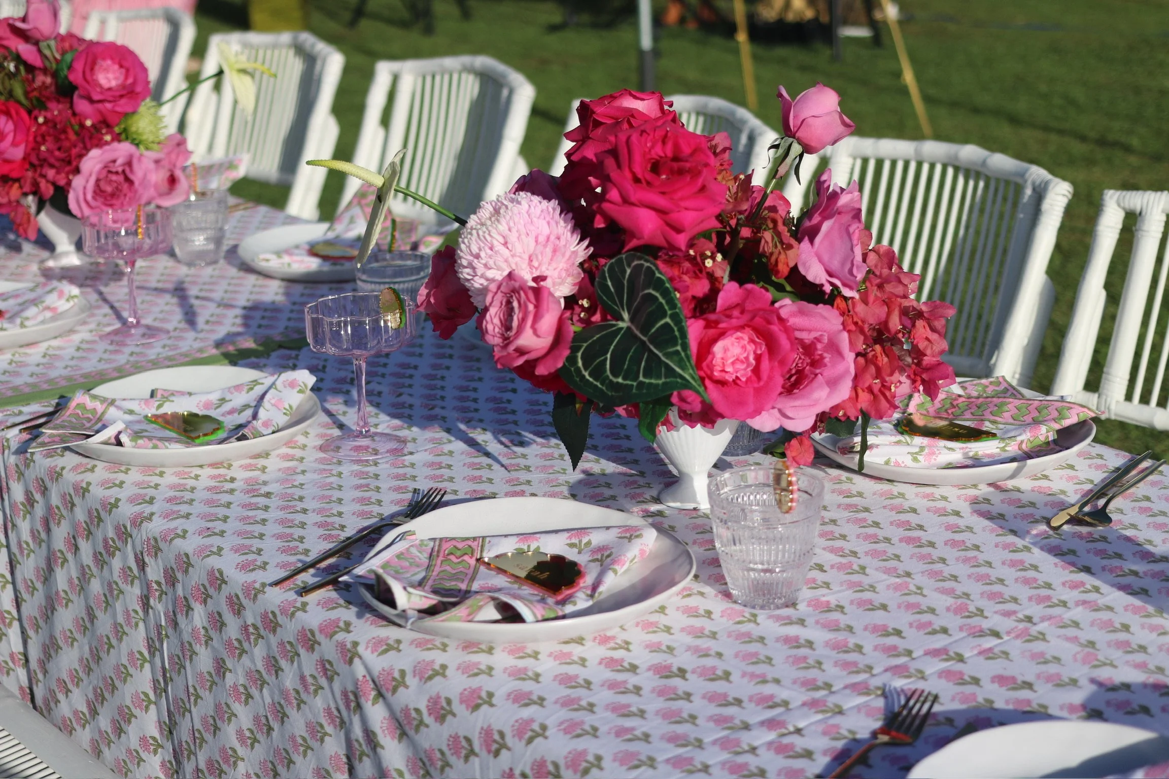 Elegant outdoor dining table set with pink and red floral centerpiece, white plates, floral napkins, cutlery, and crystal glassware, on a pink patterned tablecloth in a grassy yard.