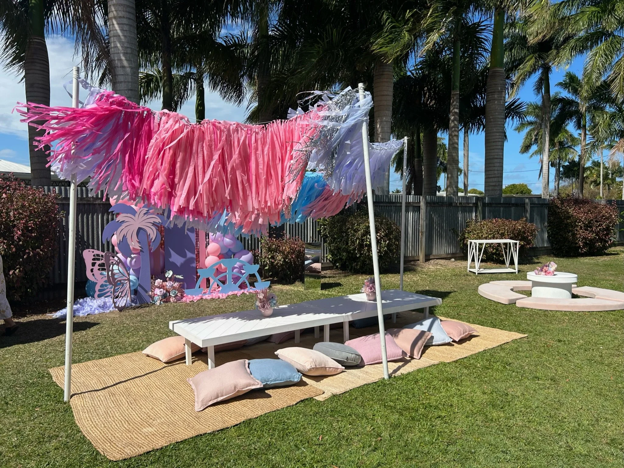 Decorative outdoor party setup with pink, purple, and white tassel garland hanging overhead, a low white table with pastel pillows and floral centerpieces on a woven rug, and a backdrop with fairy tale or fantasy theme decorations featuring a pink an
