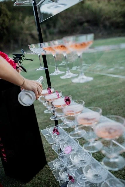 A person pouring a bottle of alcohol into a tiered display of champagne glasses outdoors.