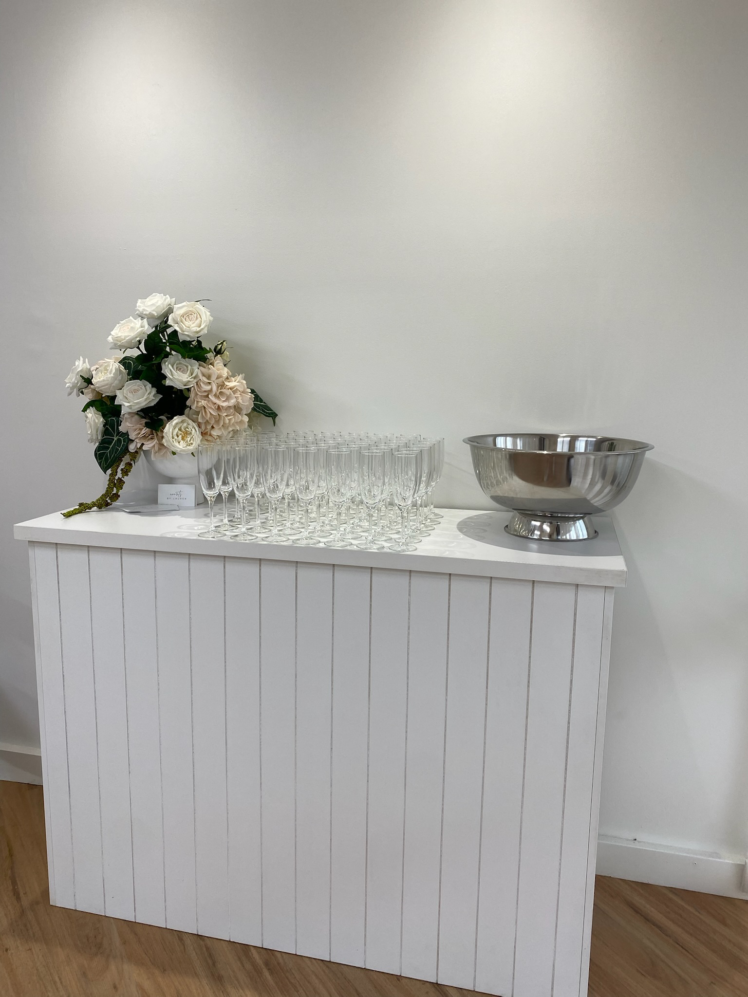 Table with a floral arrangement, rows of empty champagne glasses, and a metal bowl on a white surface in front of a white wall.