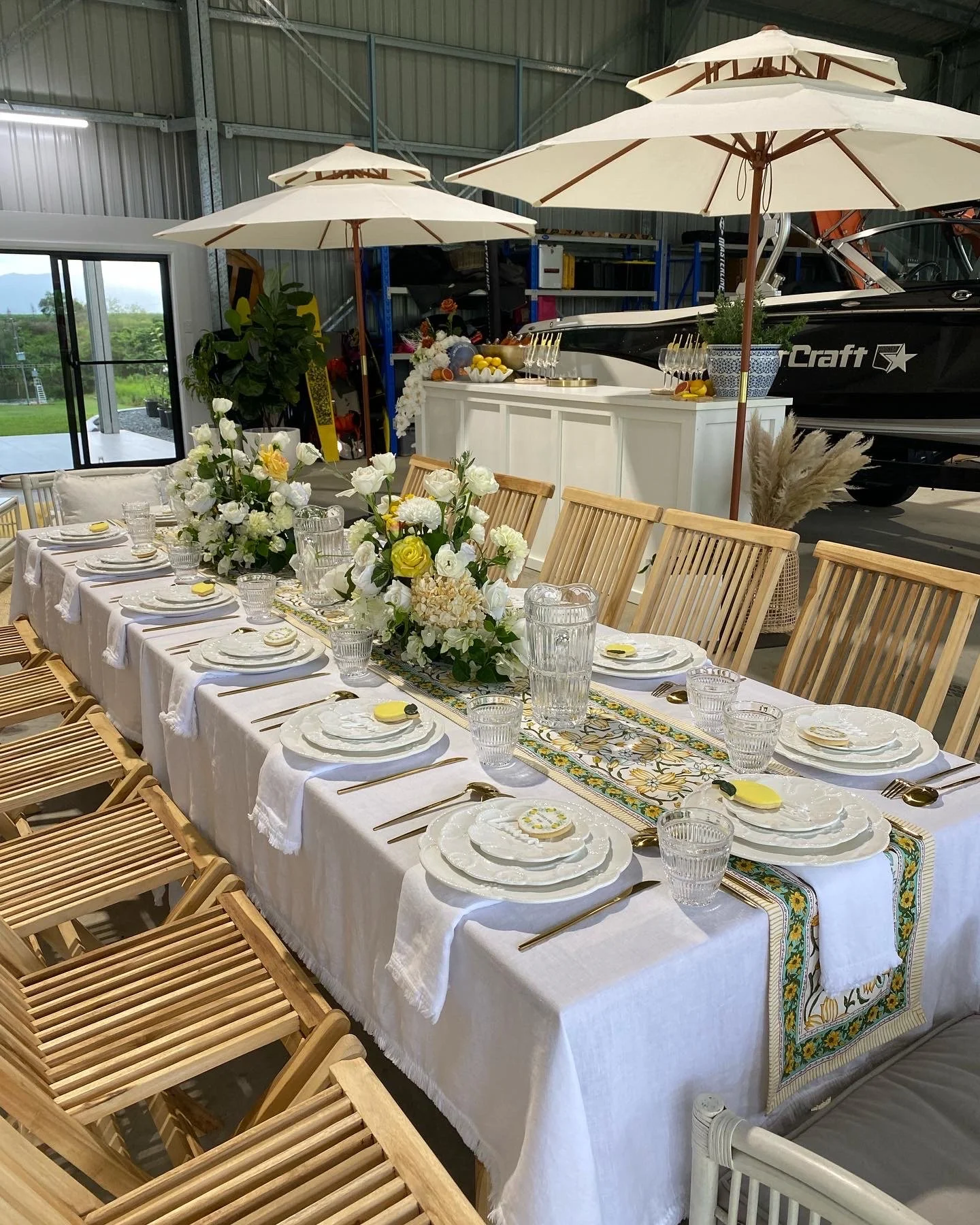A long dining table set for a gathering with white plates, glassware, gold utensils, and yellow napkins, decorated with a central floral arrangement of white and yellow flowers under two large cream umbrellas. Behind the table, there are white cabine