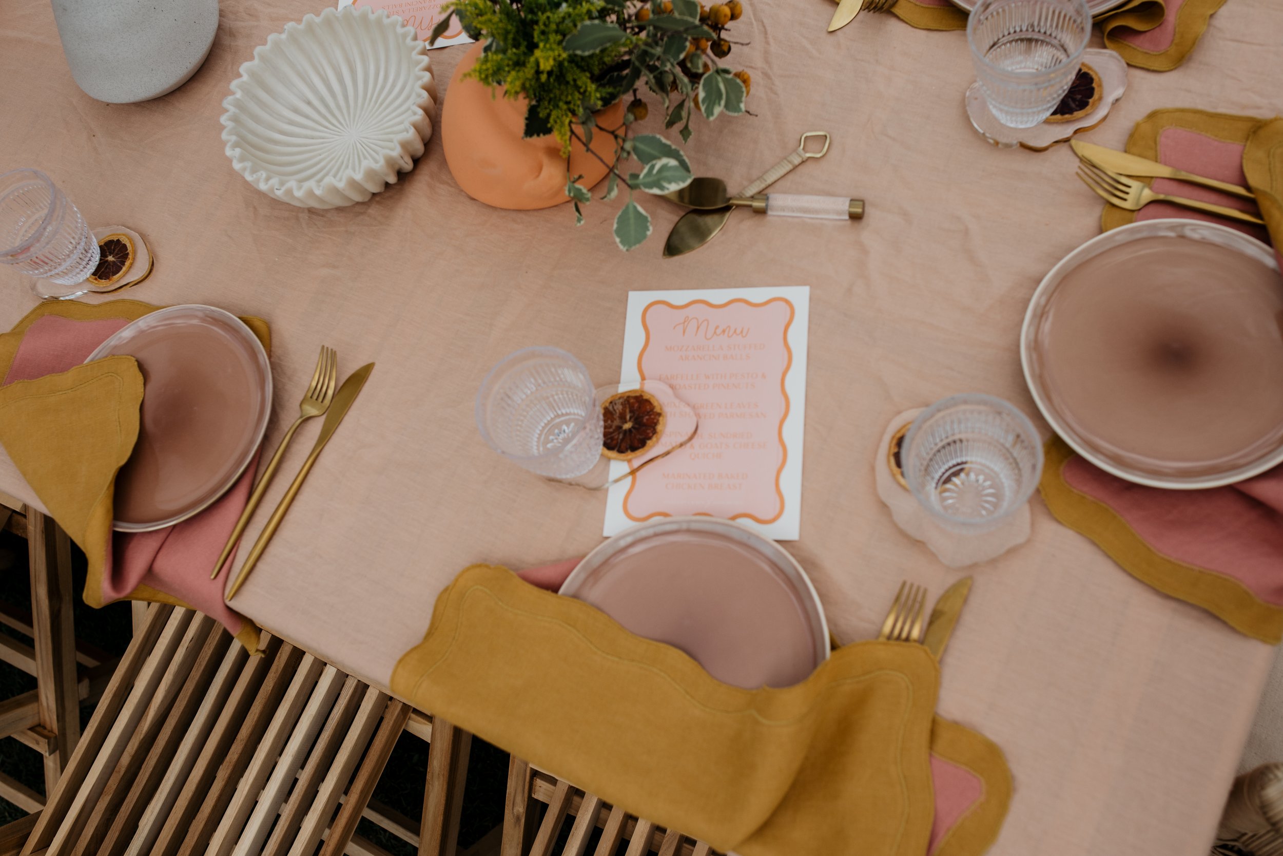 A neatly set dining table with pink and yellow napkins, gold flatware, clear glasses, and a menu in the center. There are decorative plates, a vase with green plants, and a small vase with dried flowers.