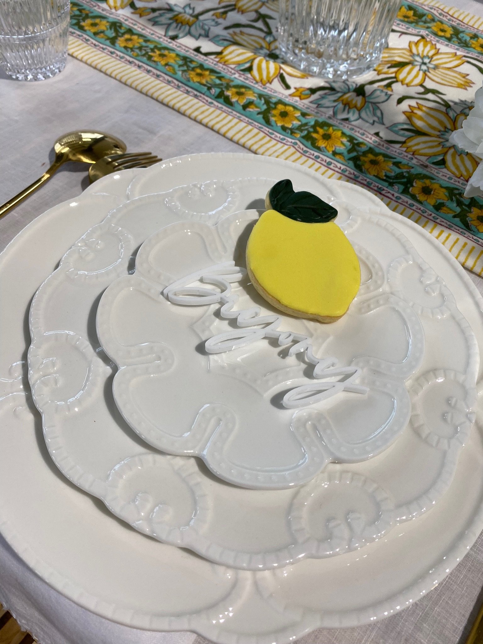 A lemon-shaped cookie with green leaves decoration on a white plate that has the word "Enjoy" in white cursive. The plate is on a table with a floral tablecloth, a glass of water, and gold-colored utensils.