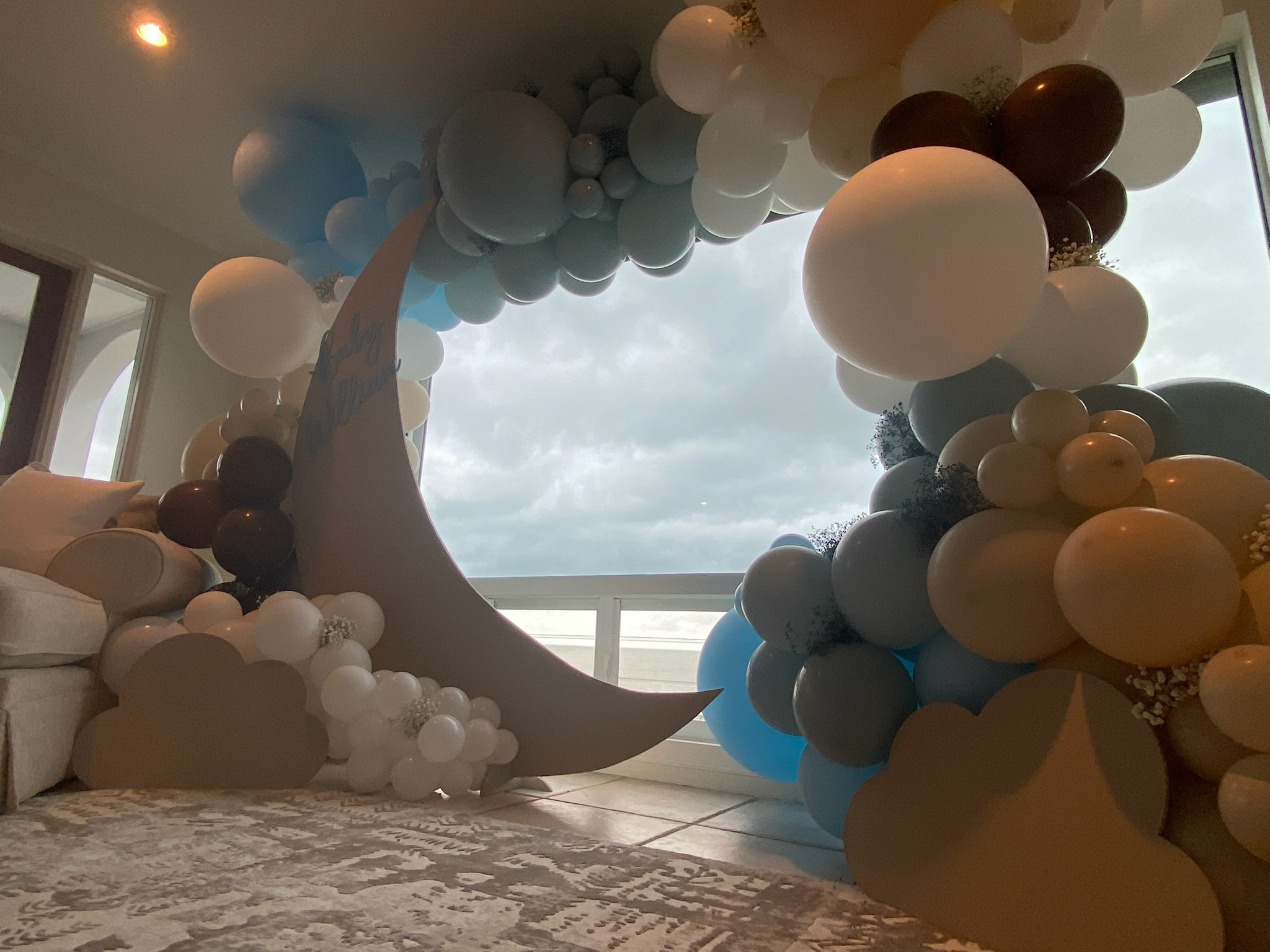 Indoor balloon decoration with a large crescent moon, clouds, and clusters of balloons in white, beige, blue, and black on a decorated area near a window with cloudy sky outside.