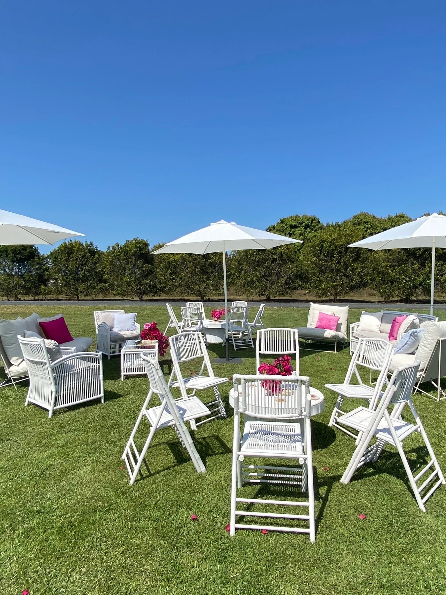 Outdoor patio setup with white chairs, sofas, and pink cushions under white umbrellas on green grass with trees in the background and blue sky overhead.