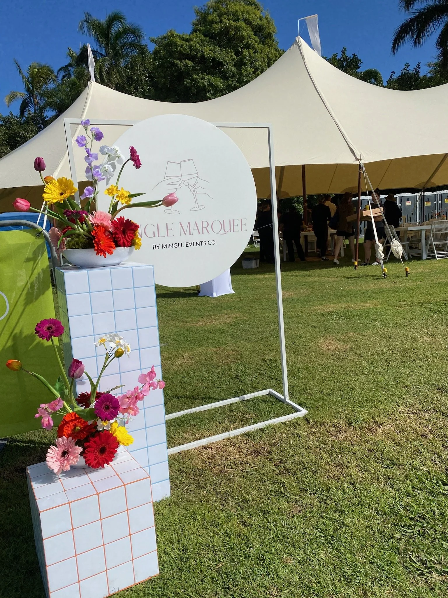 Outdoor event setup with a white tent, a sign reading 'Single Marquee by Mingle Events Co,' a display with colorful flowers, and people in the background under sunny weather.