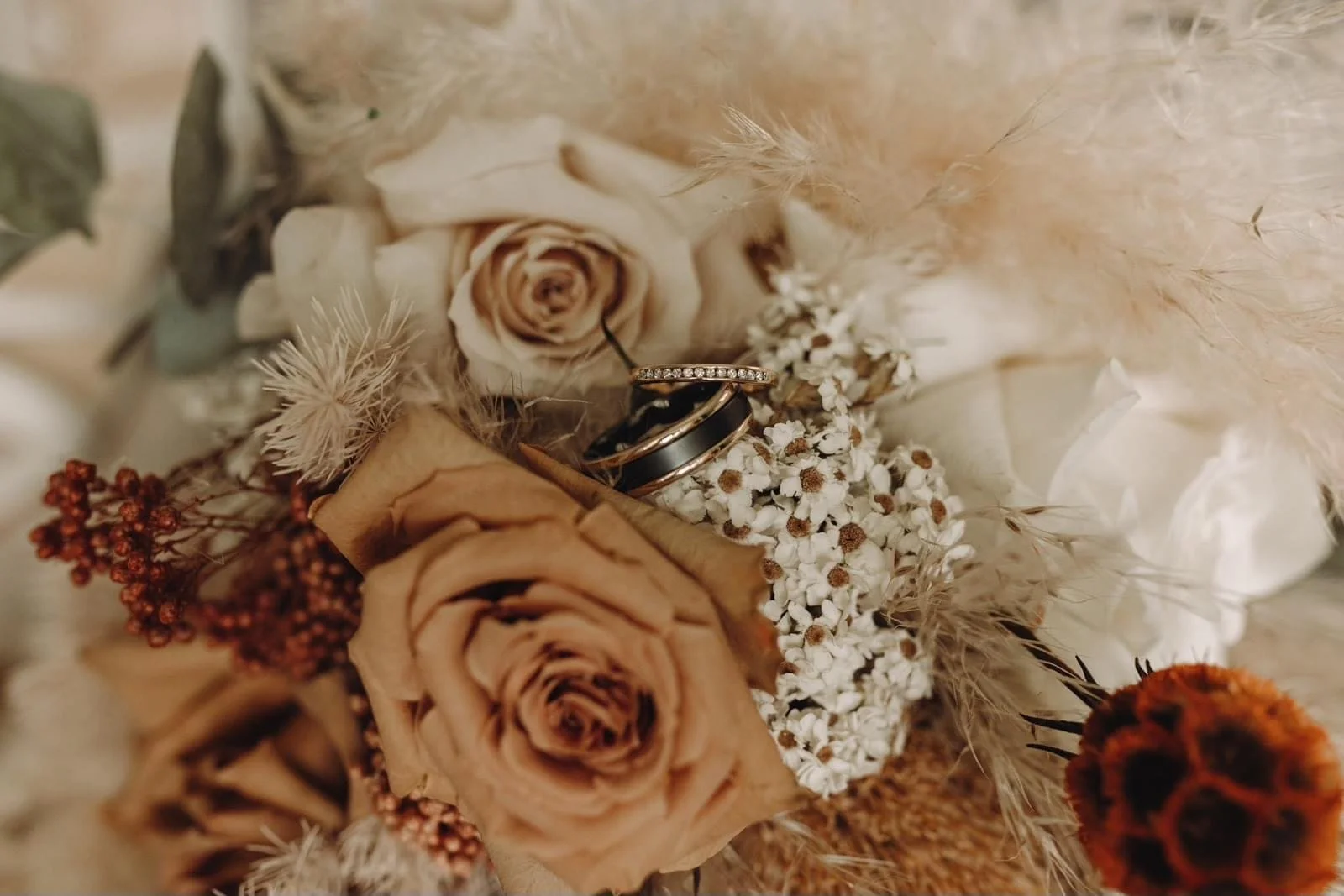 Wedding rings placed on a bouquet of dried and fresh flowers, including beige roses, white baby's breath, and dried grasses.
