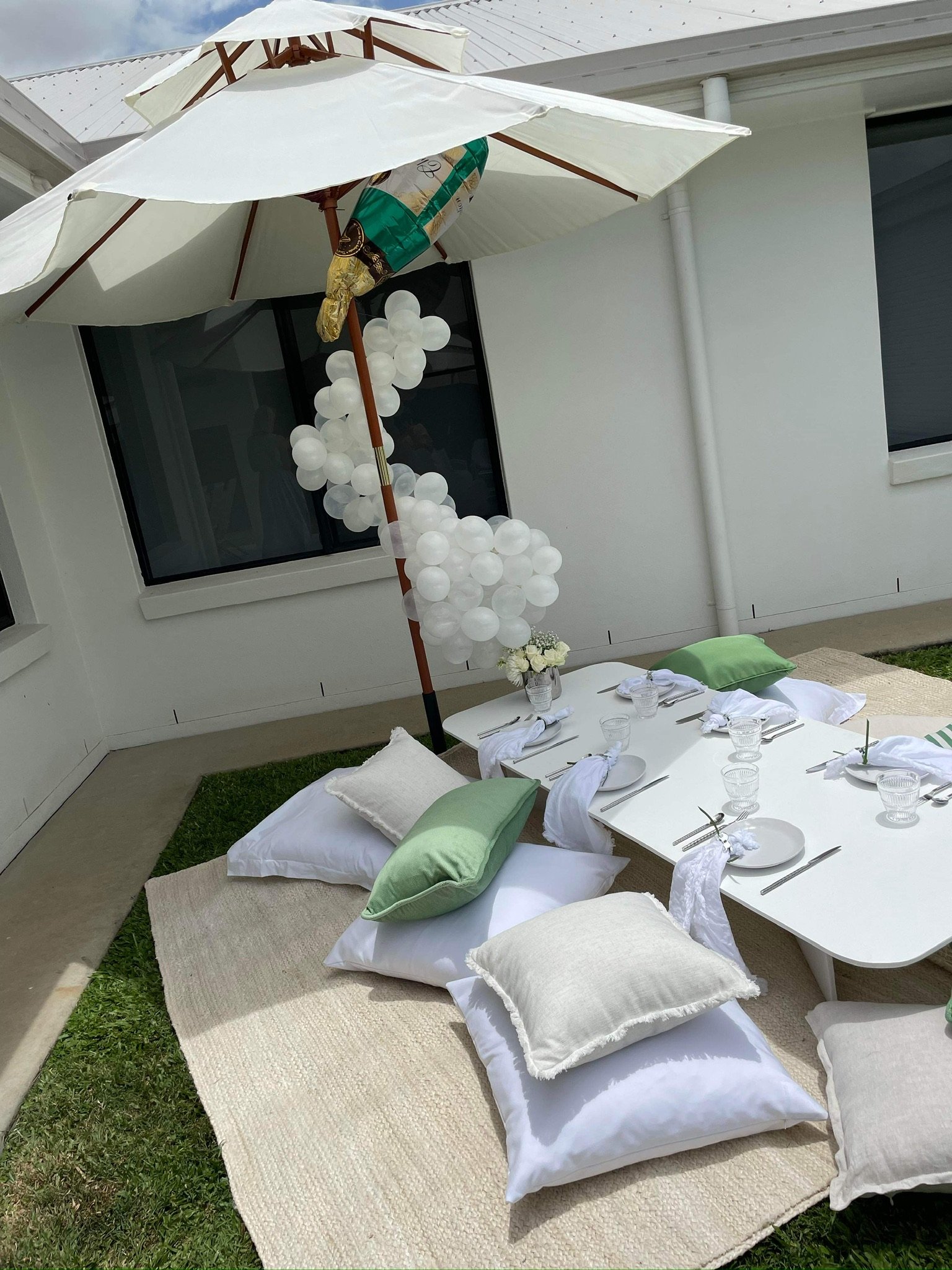 Outdoor dining setup with a white table, pillows on the ground, a large white umbrella, white balloons arranged in a cluster, and a vase of white flowers
