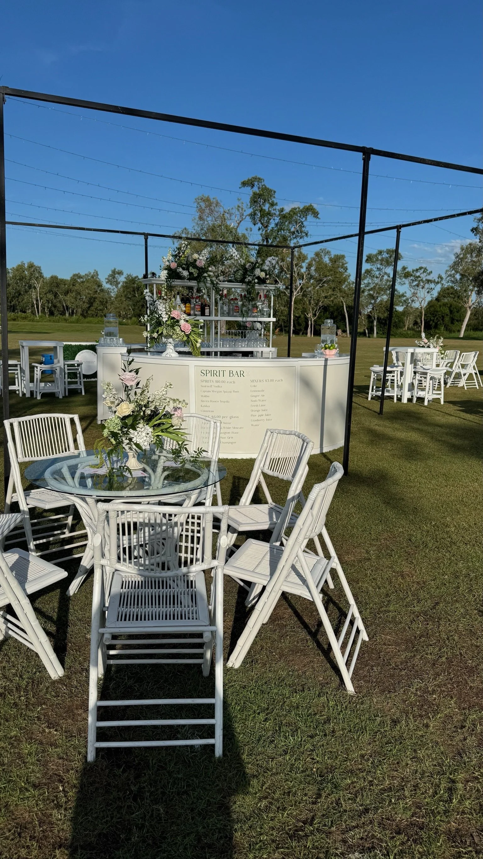 Outdoor event setup with round glass table and white chairs, a white bar labeled 'Spirit Bar' with floral decorations, against a grassy field and blue sky.