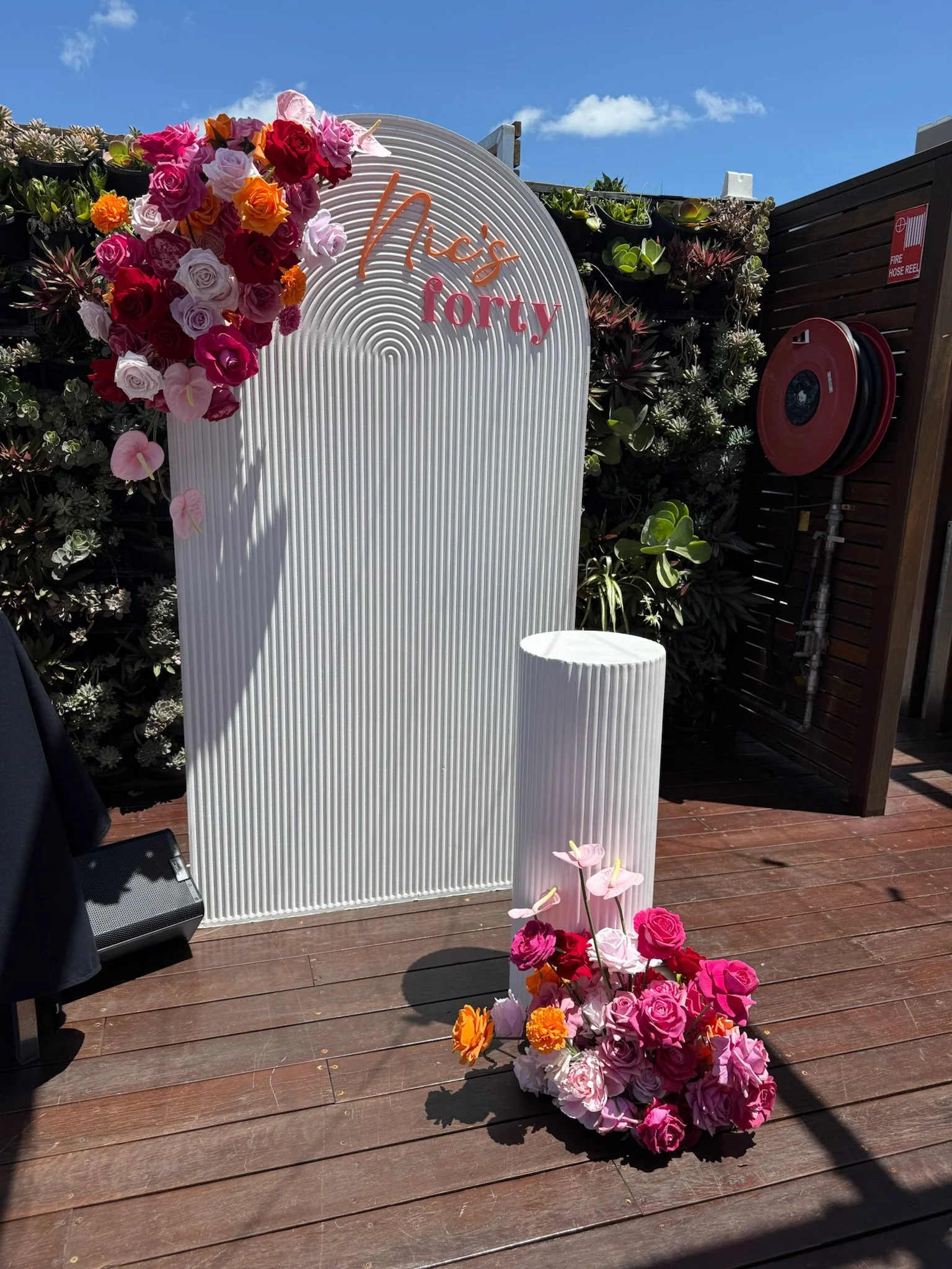 Decorative backdrop with flowers and the words "Mrs. Tori" on it, featuring a large white arched panel and a smaller cylindrical pedestal, both adorned with colorful pink, red, orange, and white flowers.