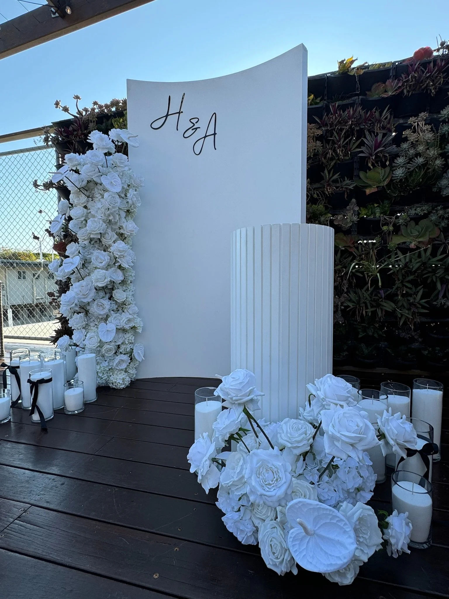 White floral arrangement with candles and a curved white backdrop with the initials H&A, set on a wooden floor outdoors.