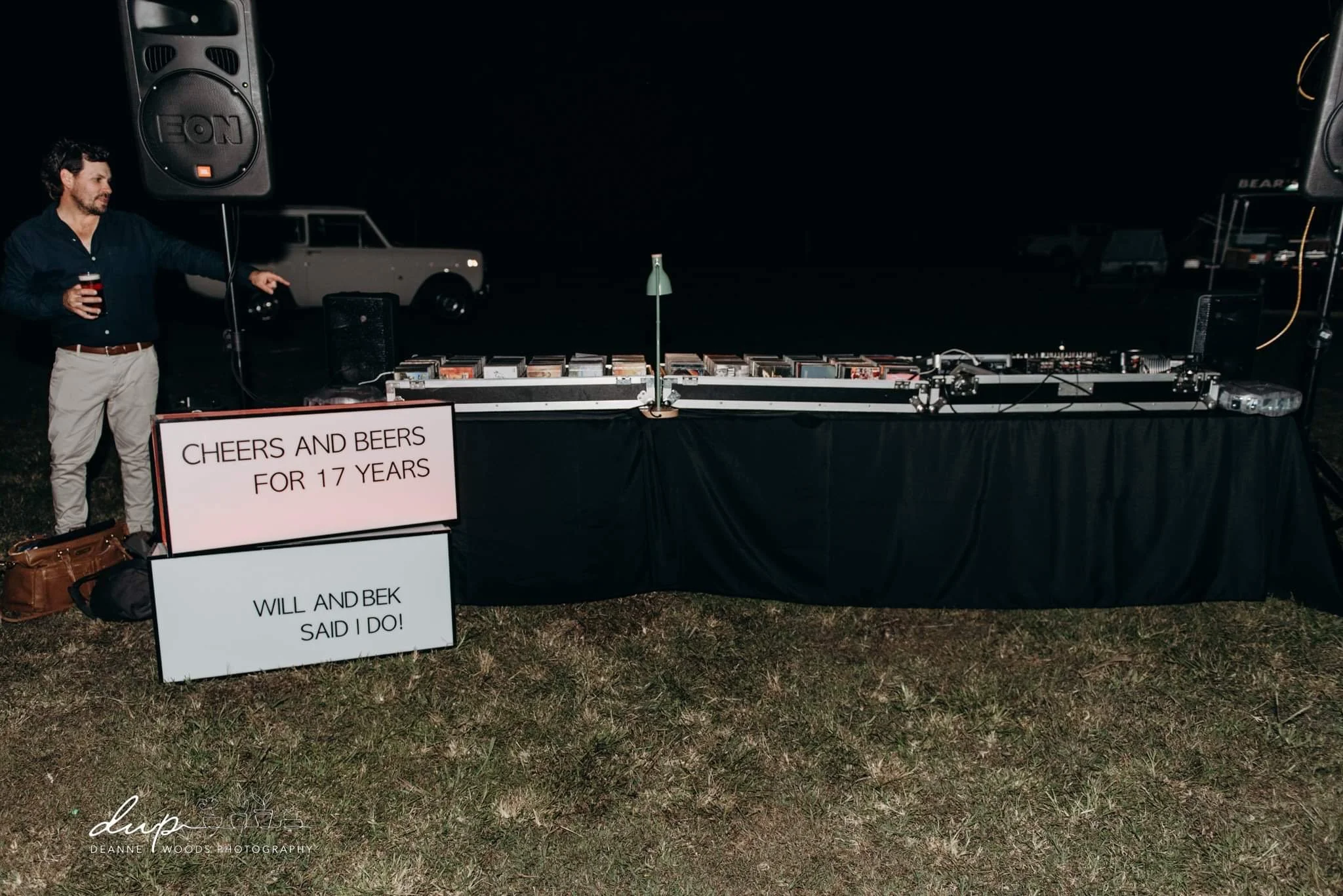 Outdoor DJ setup at night with a man holding a drink pointing at the equipment, two signs reading 'Cheers and Beers for 17 Years' and 'Will and Bek Said I Do!', black tablecloth, speakers, and a vintage truck in the background.