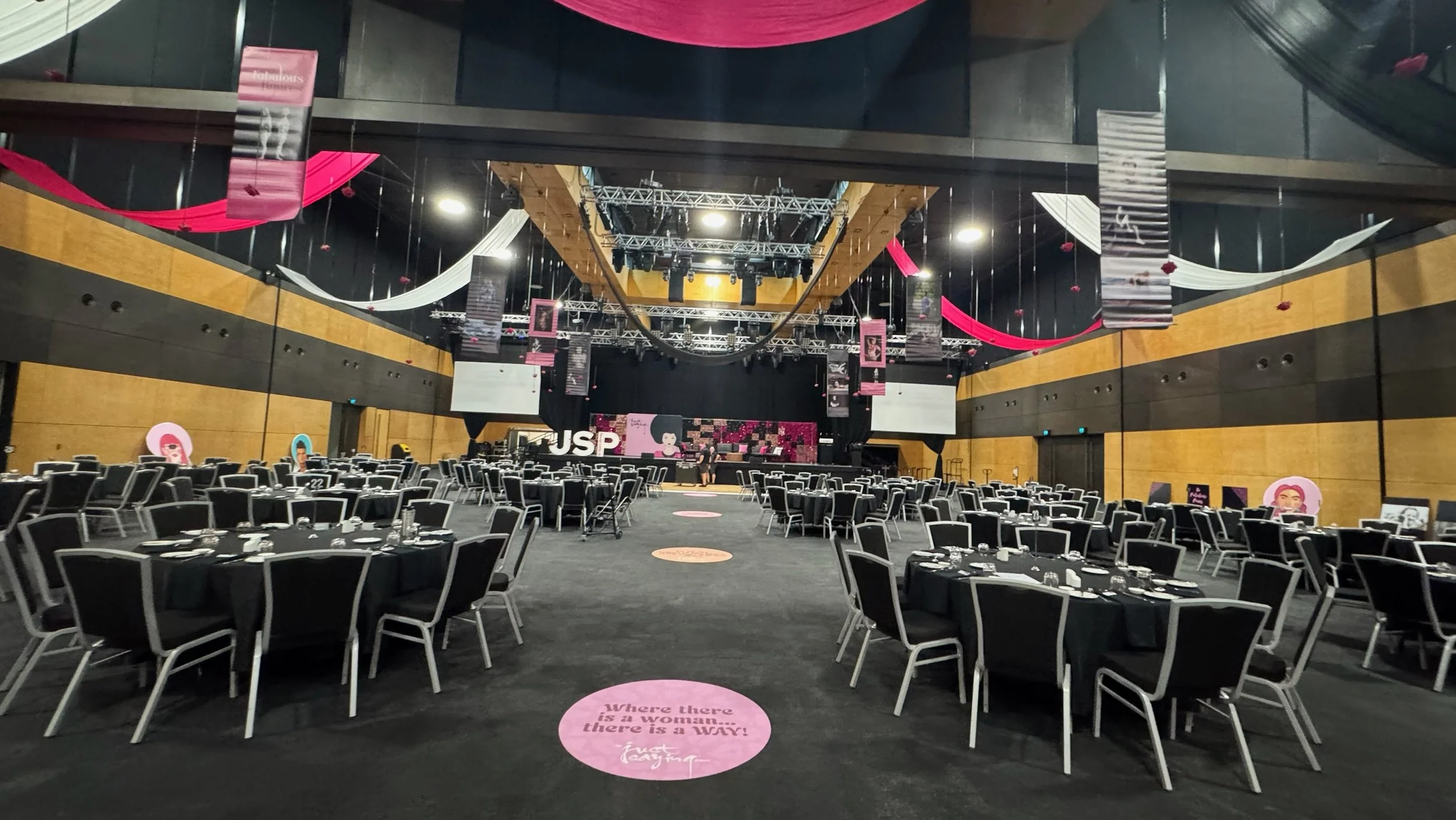 Empty event hall with round tables covered in black tablecloths, set for a formal event, with a stage at the front and pink and white decorations hanging from the ceiling.