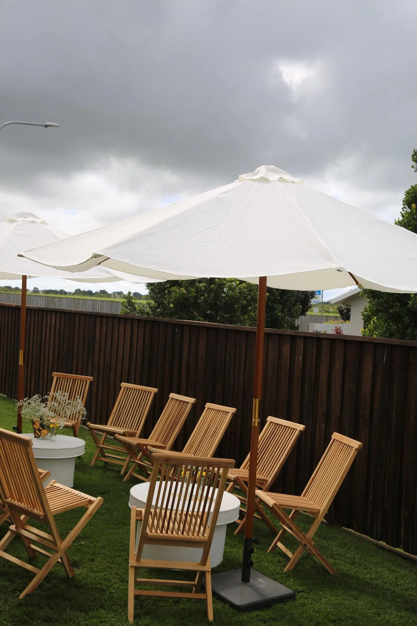 Outdoor patio area with wooden chairs, small tables, and large white umbrellas under a cloudy sky.