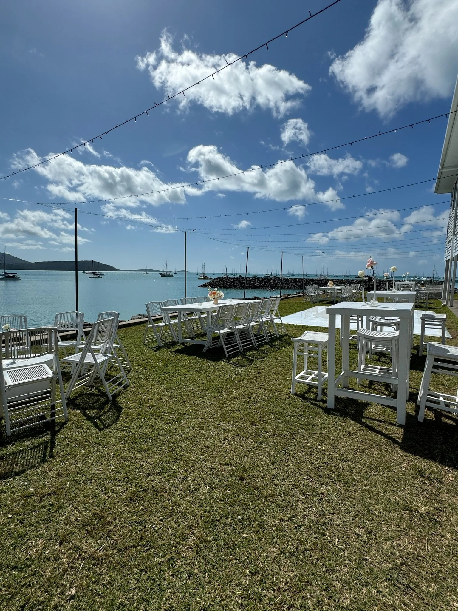 Outdoor waterfront seating area with white tables and chairs, string lights overhead, and boats on the water under a partly cloudy blue sky.