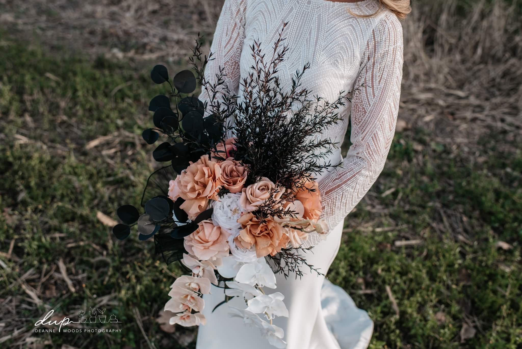 A woman in a white lace dress holding a bouquet of peach, white, and blush roses, black and green foliage, standing outdoors on grass.
