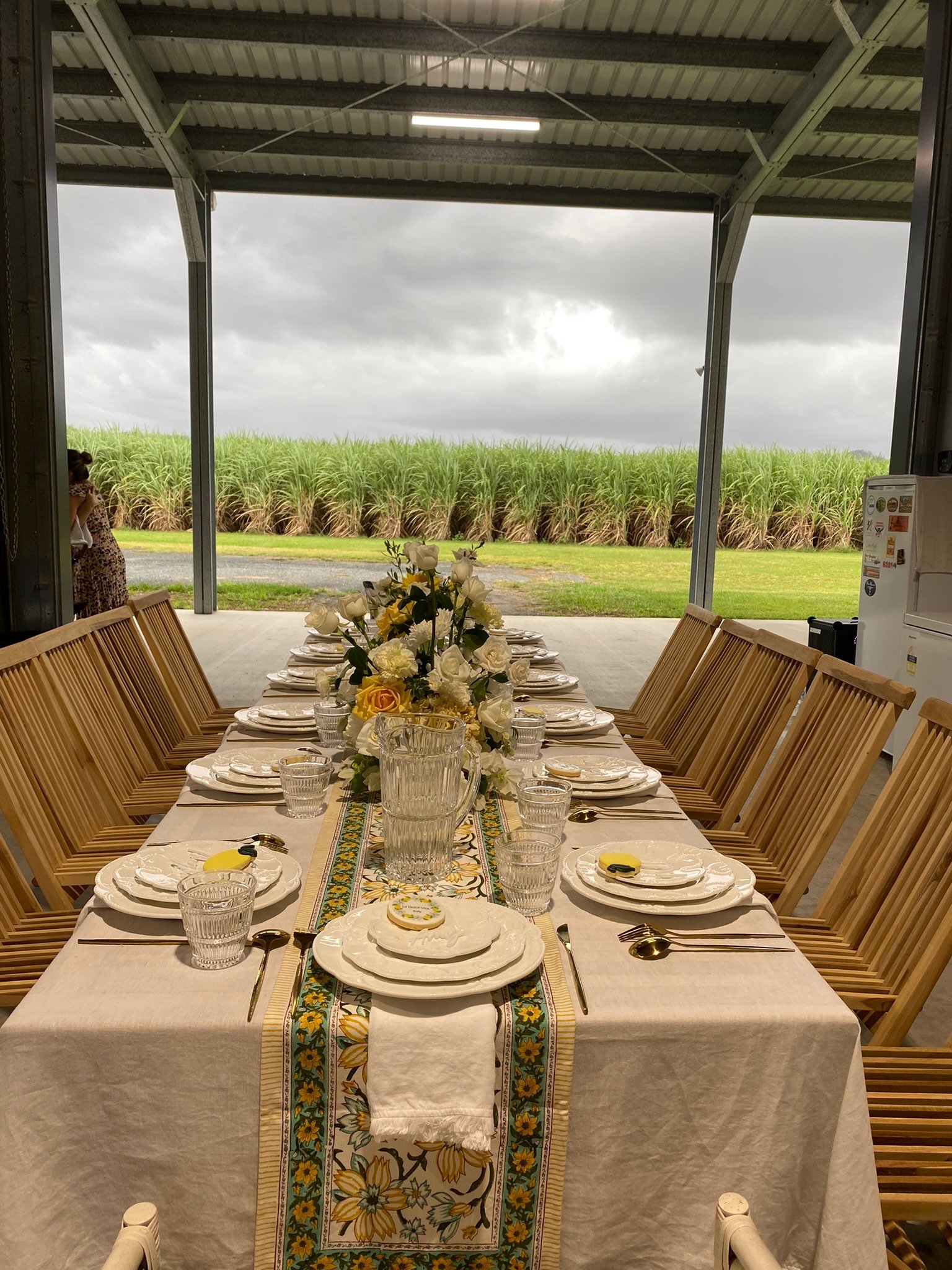 Long dining table set with white plates, cutlery, water glasses, yellow and white floral centerpiece, and a decorative table runner with sunflower pattern. The table is in a covered outdoor area overlooking a field of sugarcane, with a cloudy sky.