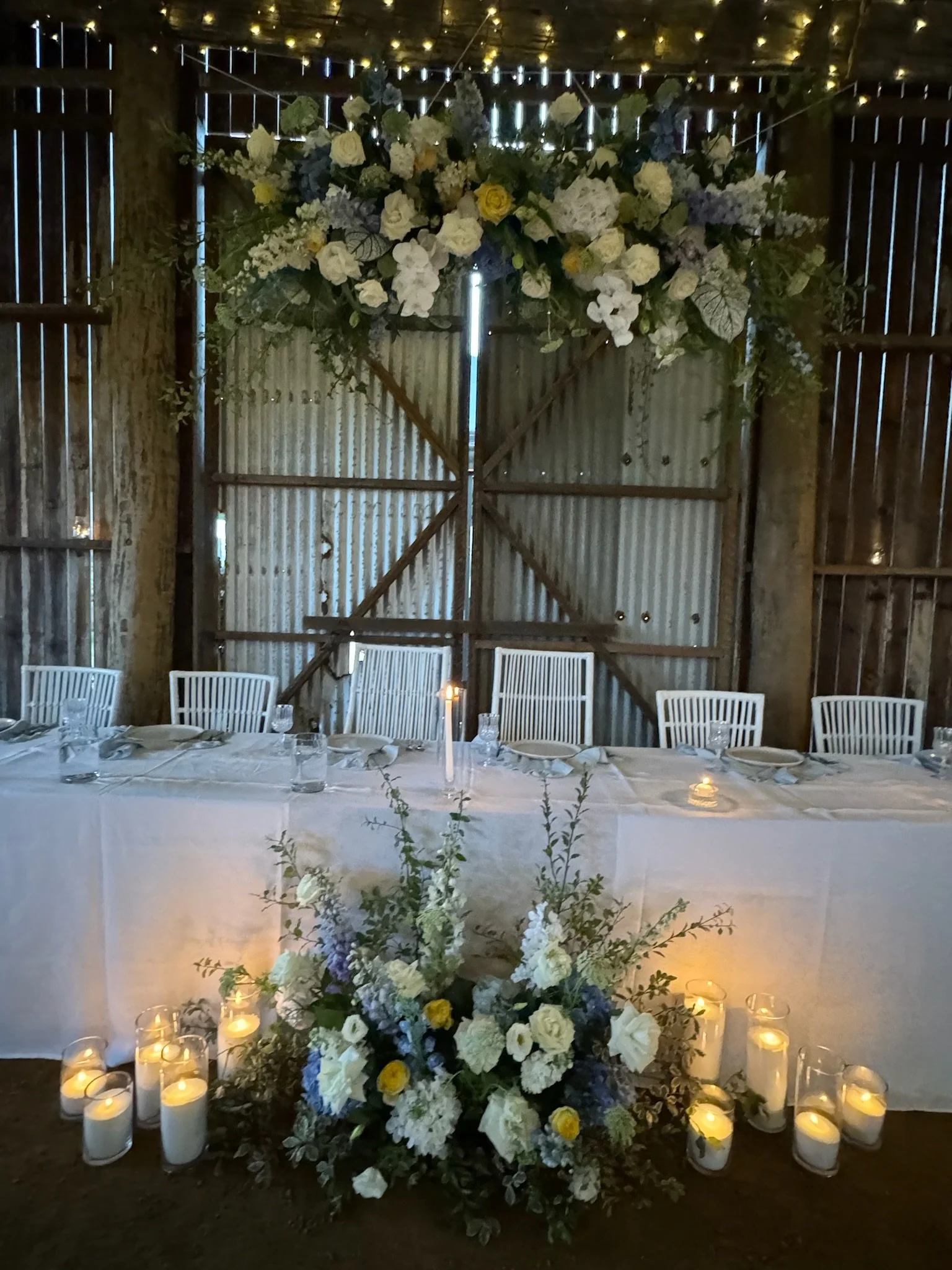 Rustic barn interior decorated for a wedding with a long table featuring a floral centerpiece, candles, and a large floral arrangement hanging above.