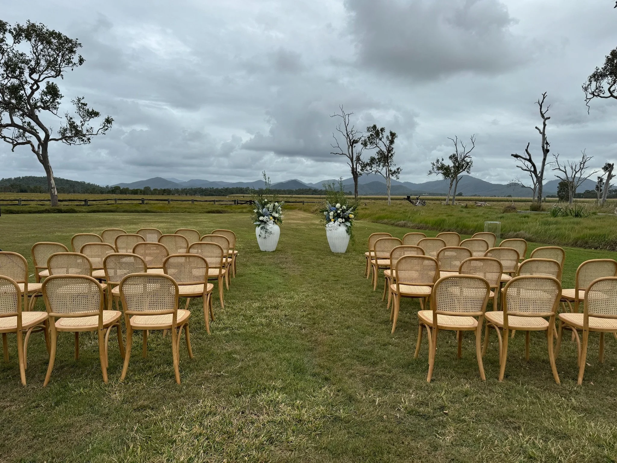 An outdoor wedding setup with rows of wooden chairs facing two large white vases with white and green floral arrangements, set on a grassy field with trees, mountains, and cloudy sky in the background.