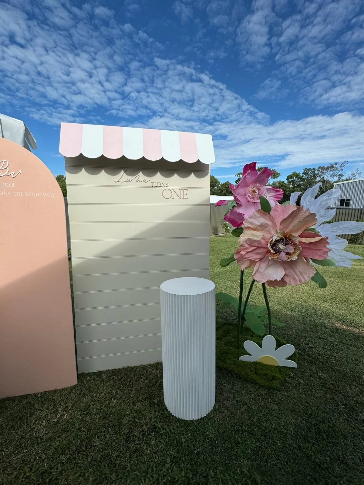 Outdoor display with large artificial pink and white flowers, a white cylindrical stand, and a white board with pink and white striped awning, against a blue sky with scattered clouds.