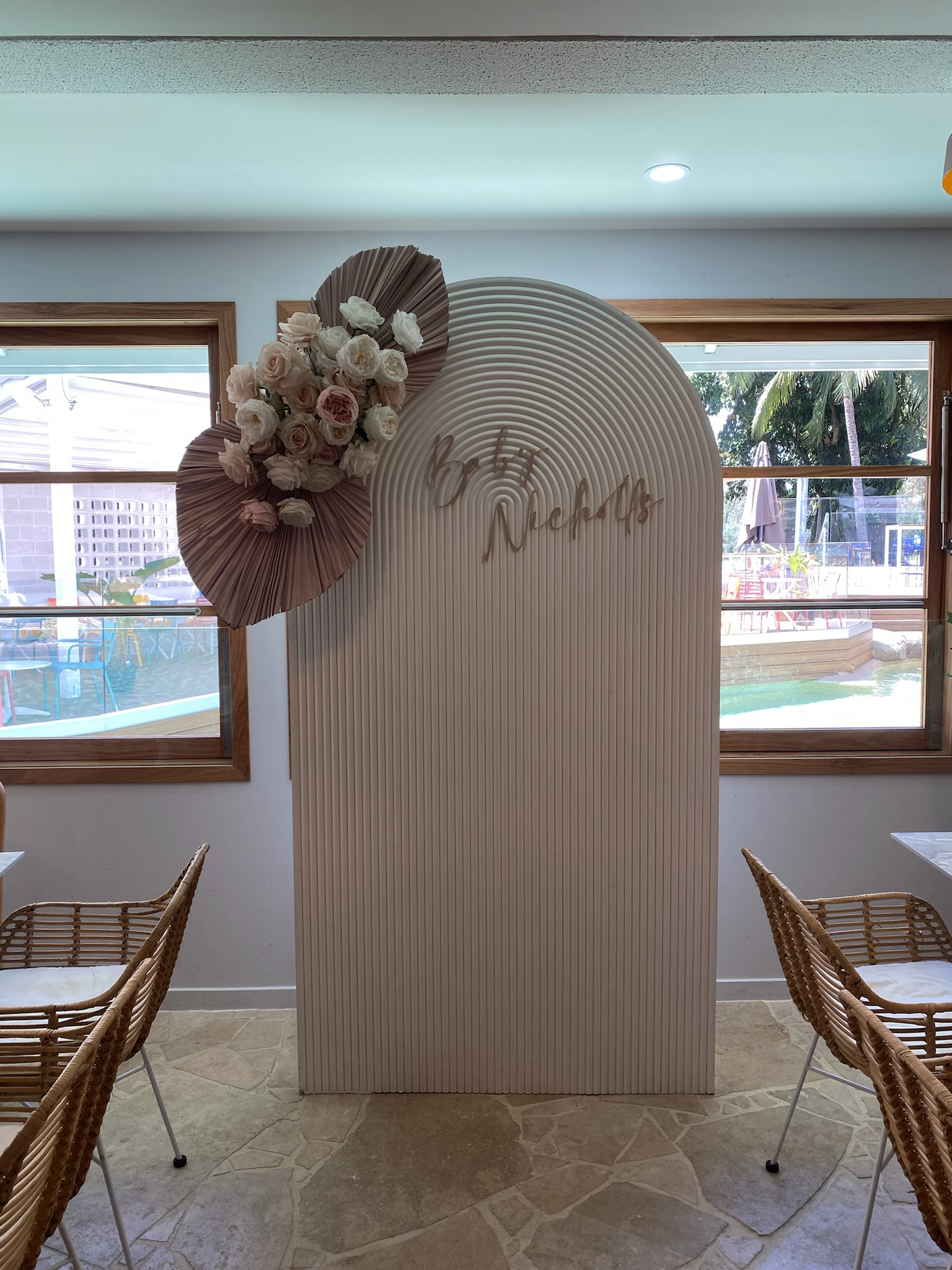 Decorative backdrop with white and pink flowers, pleated design, and script sign reading 'The Rickeys', in a restaurant or cafe with windows and wicker chairs.