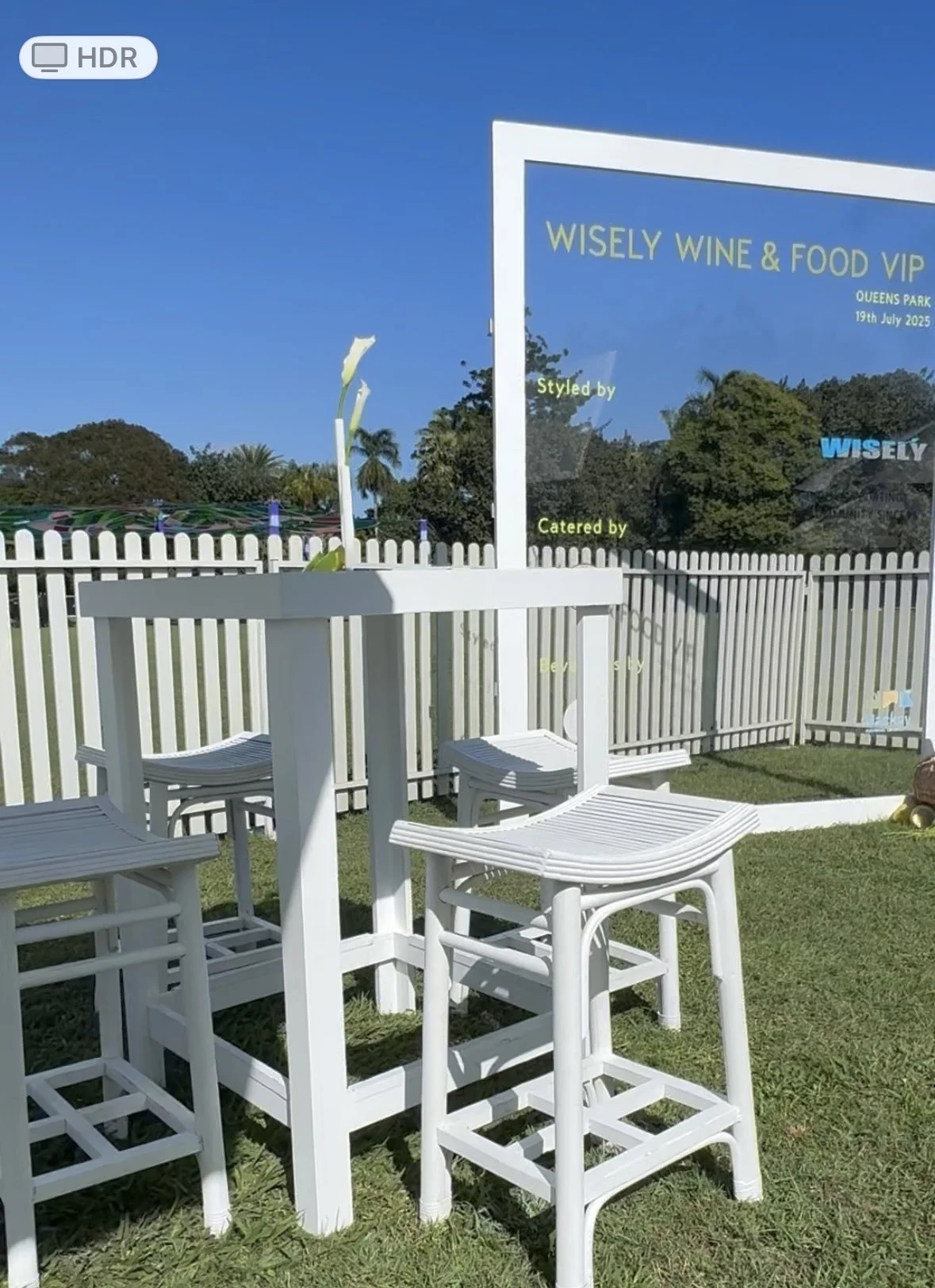 White bar table with three white bar stools on green grass, enclosed by a white picket fence, with a sign that reads 'WISELY WINE & FOOD VIP' in the background.