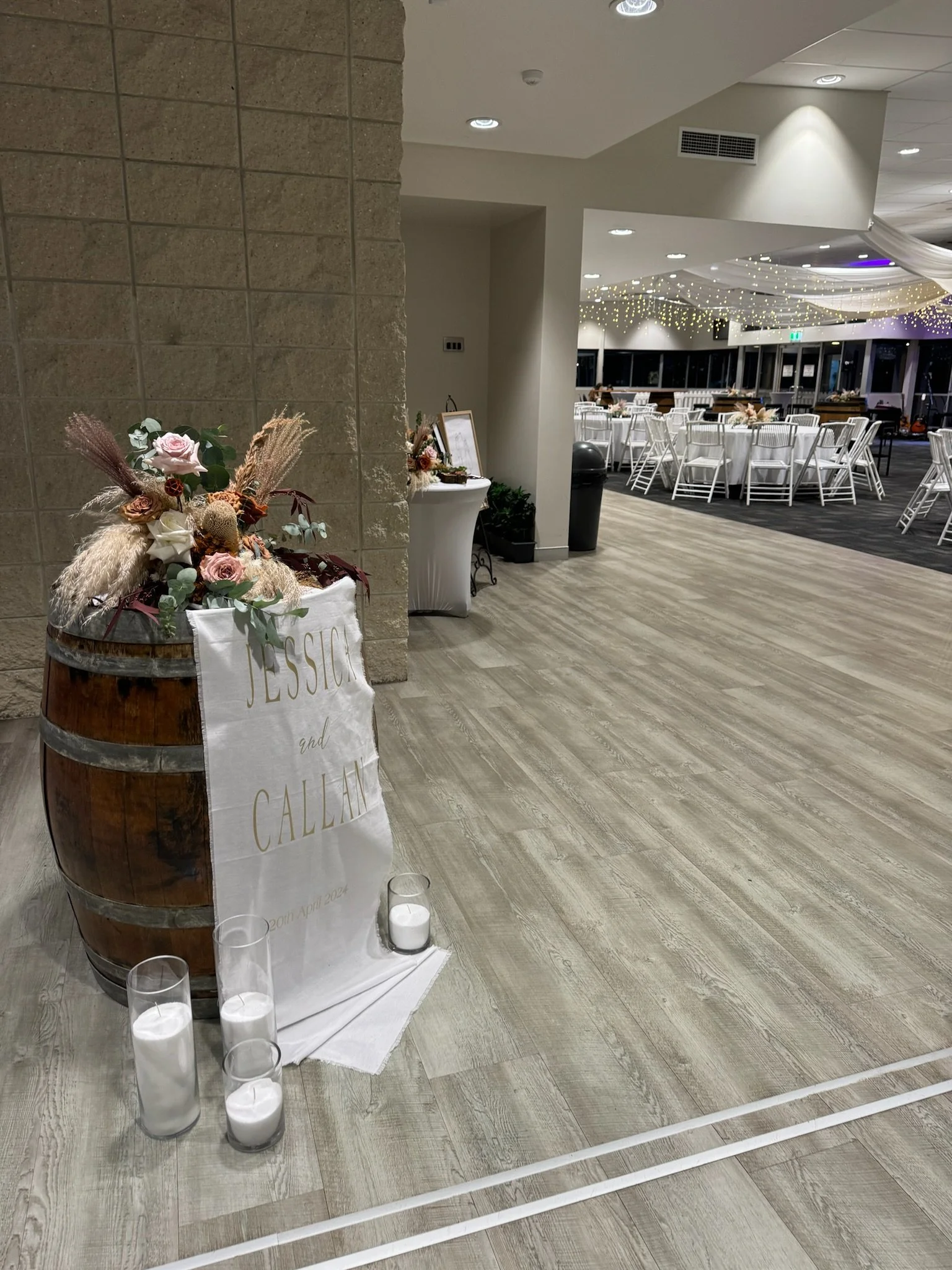 Decorative display with flowers, candles, and a sign at a wedding reception venue, with tables and chairs visible in the background.