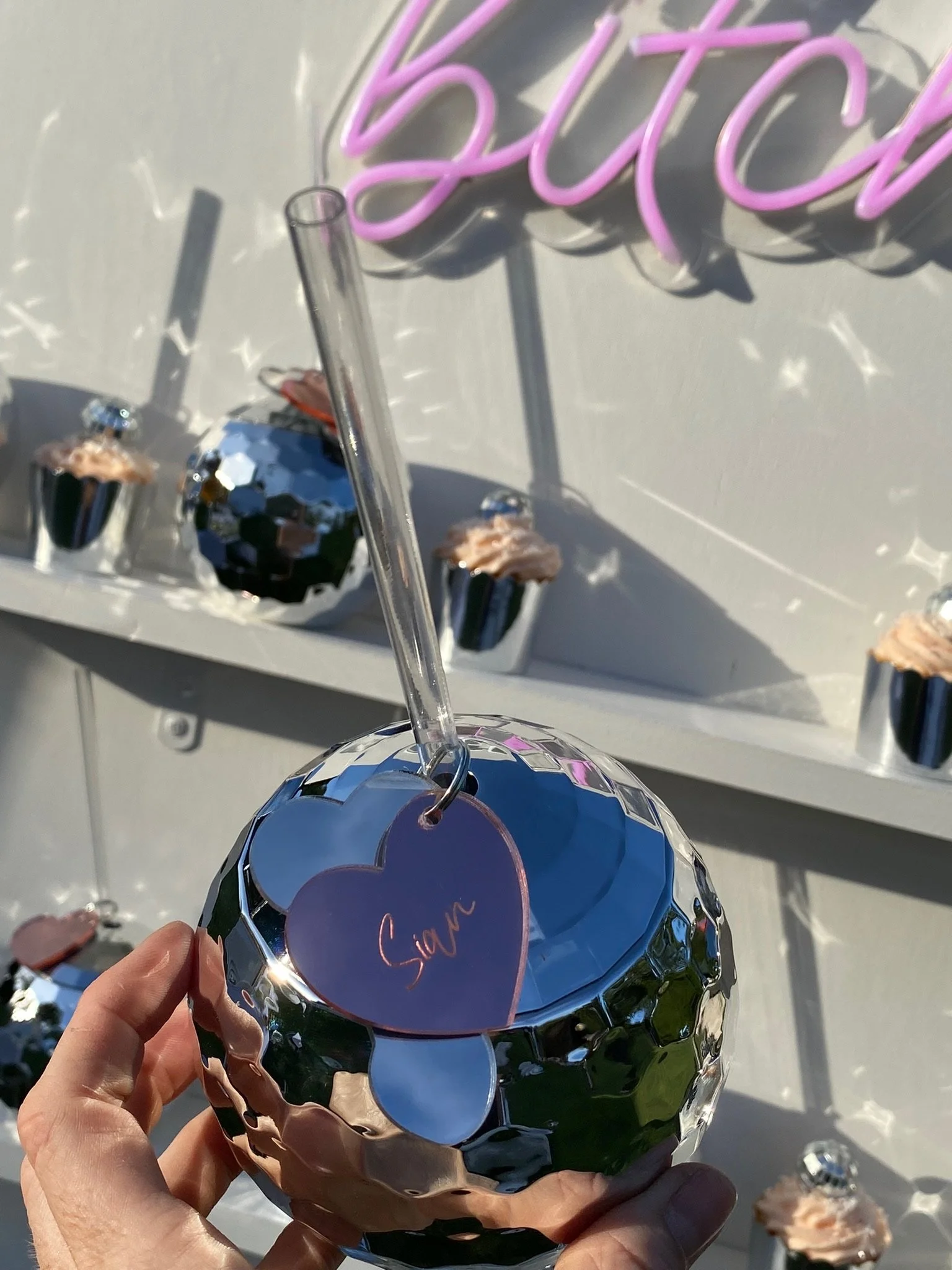 A hand holding a small silver disco ball with a purple heart-shaped tag that says 'Sian' attached to a straw, with cupcakes in silver wrappers on a white shelf behind it and a pink neon sign reading 'butch' in the background.