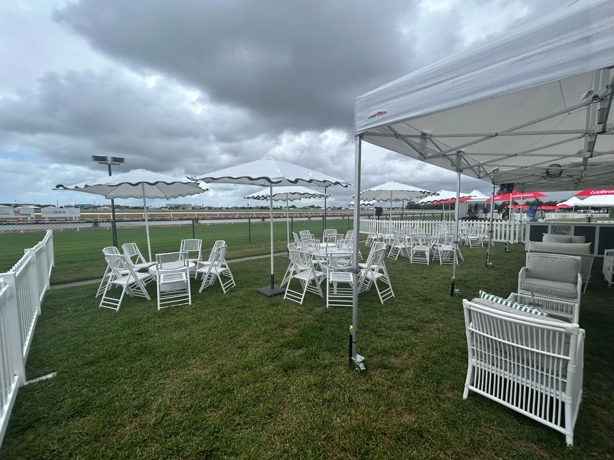 Empty outdoor seating area with white chairs and tables under white umbrellas, fenced area with a grassy field and cloudy sky.