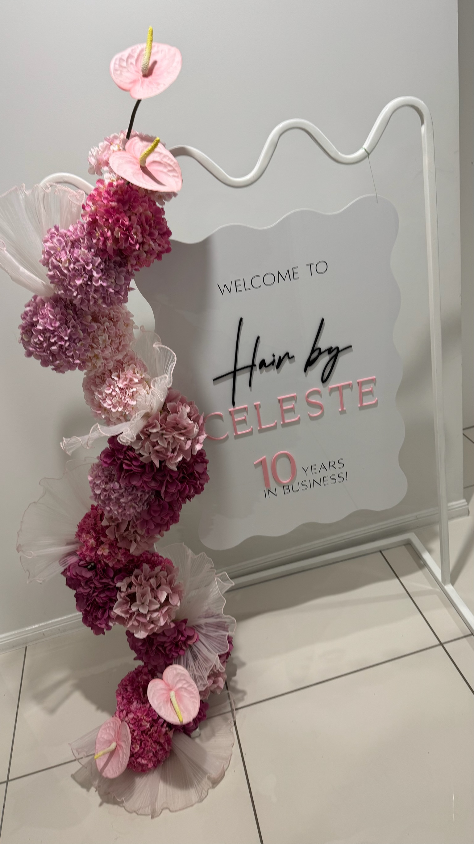 Decorative sign welcoming clients to Hair by Celeste celebrating 10 years in business, adorned with pink flowers and an anthurium floral arrangement.