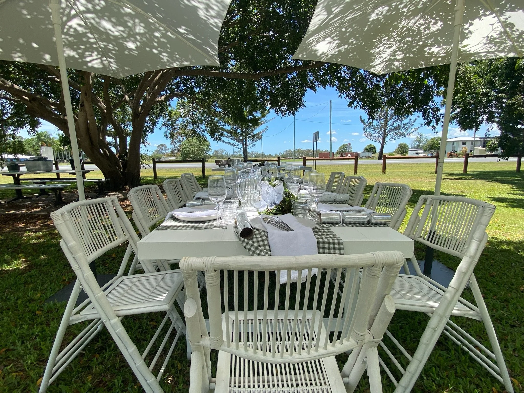 A long outdoor dining table under a shaded tree with a checkered table runner, set with white plates, silverware, water glasses, and wine glasses, surrounded by white chairs with umbrellas providing shade, on a grassy area with a clear blue sky in th