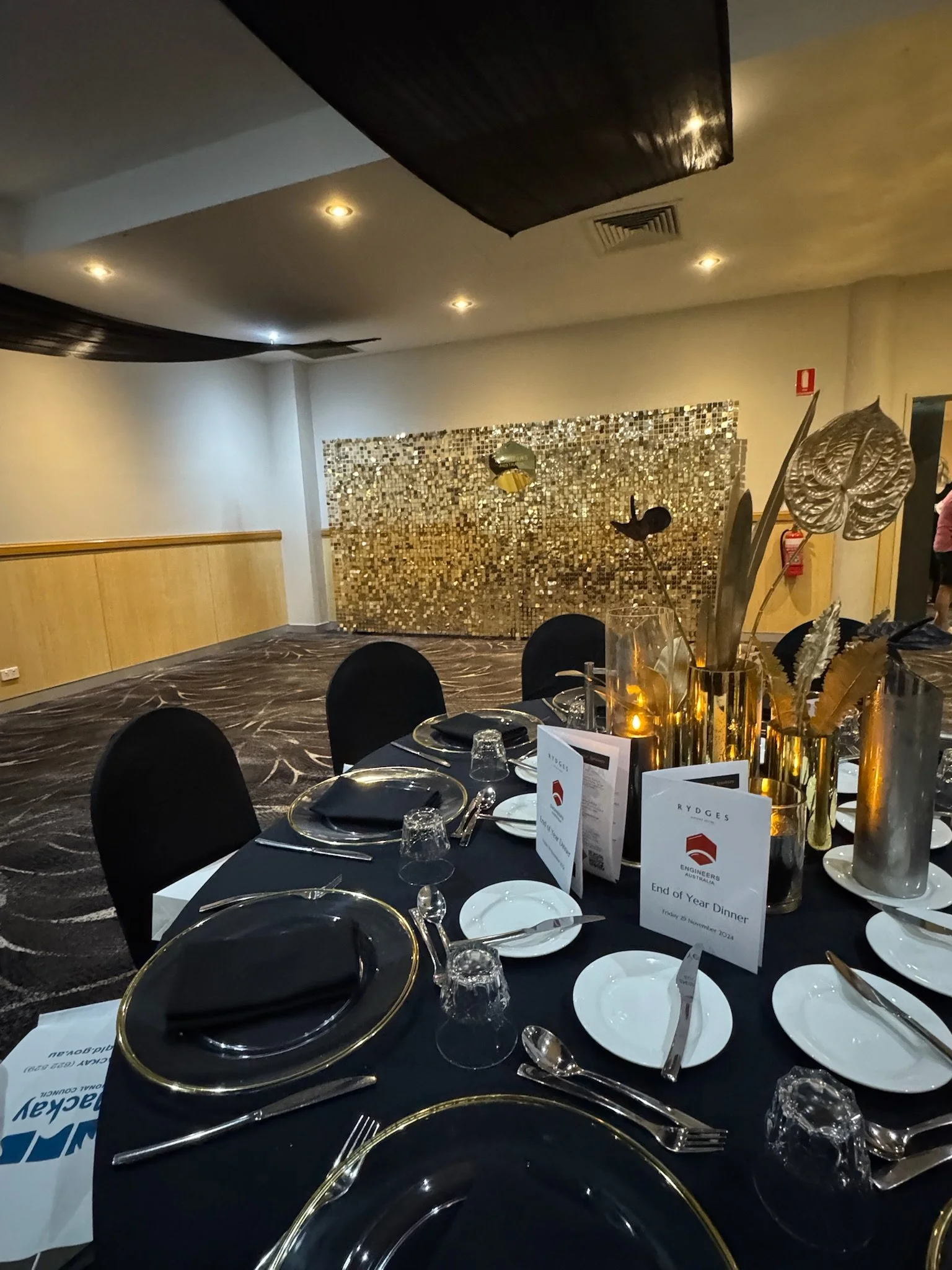 A formal dining table set for a dinner event with black tablecloths, black napkins, and gold accents. The table has white plates, silverware, wine glasses, and a tall gold vase with decorative metallic leaves and flowers. In the background, there is 