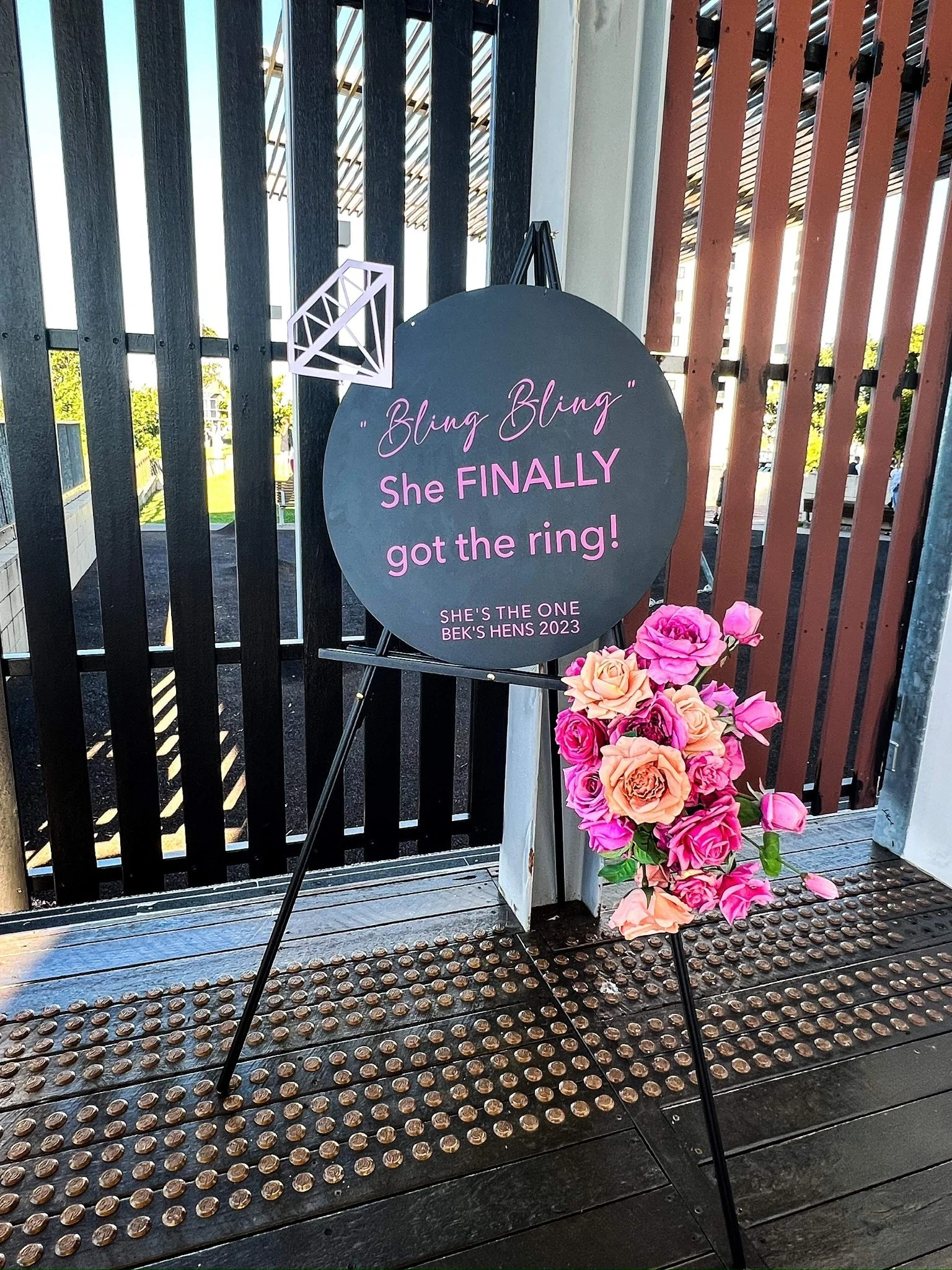 A decorative sign with pink and purple lettering on a black circular background, placed on a black easel. The sign reads, "Bling Bling she FINALLY got the ring!" with additional text at the bottom stating, "SHE'S THE ONE BEK'S HENS 2023." Next to the