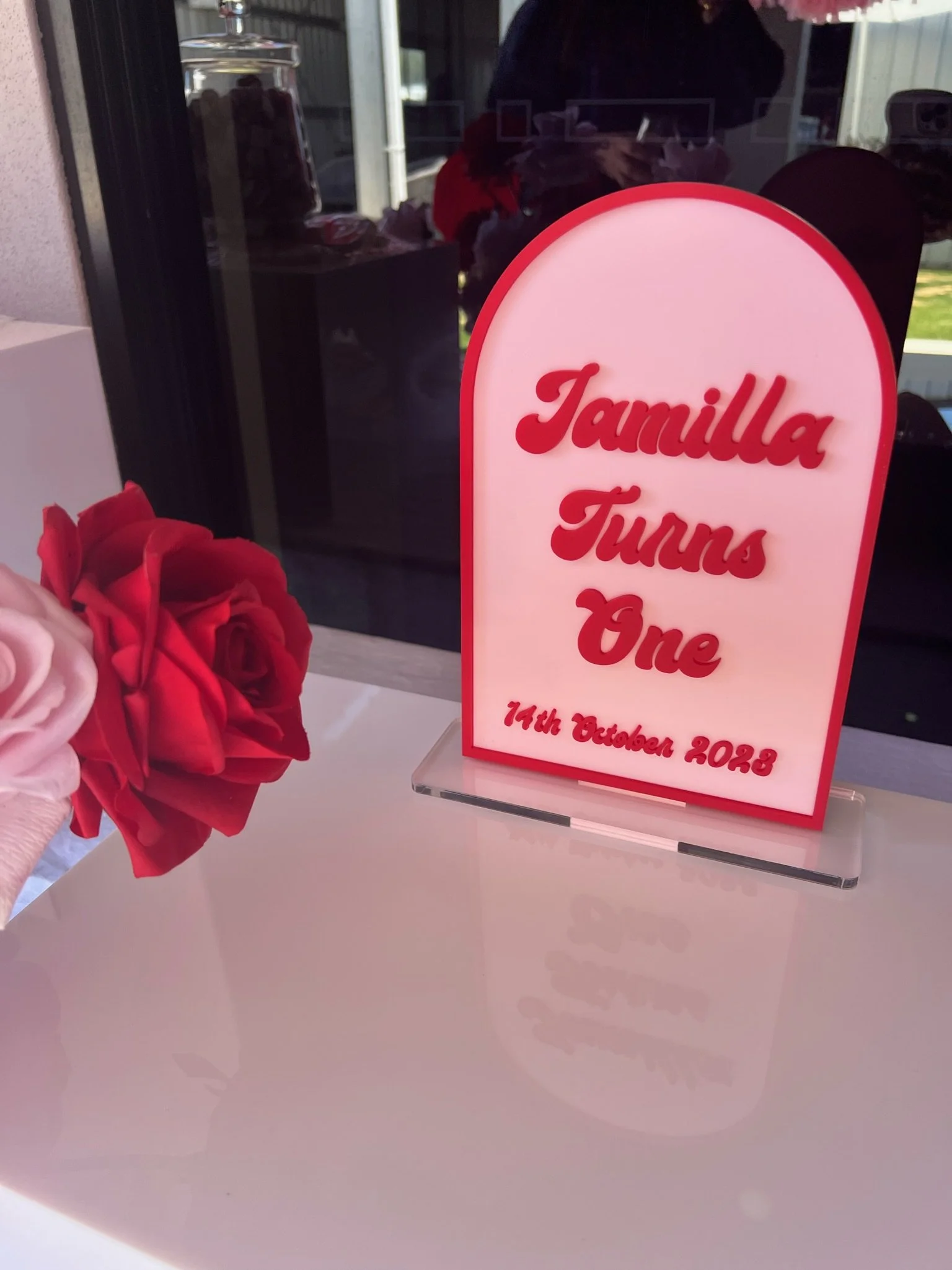 A digital sign celebrating a family birthday with the text 'Tamilla Turns One, 14th October 2023,' placed on a white table next to a bouquet of pink and red roses.