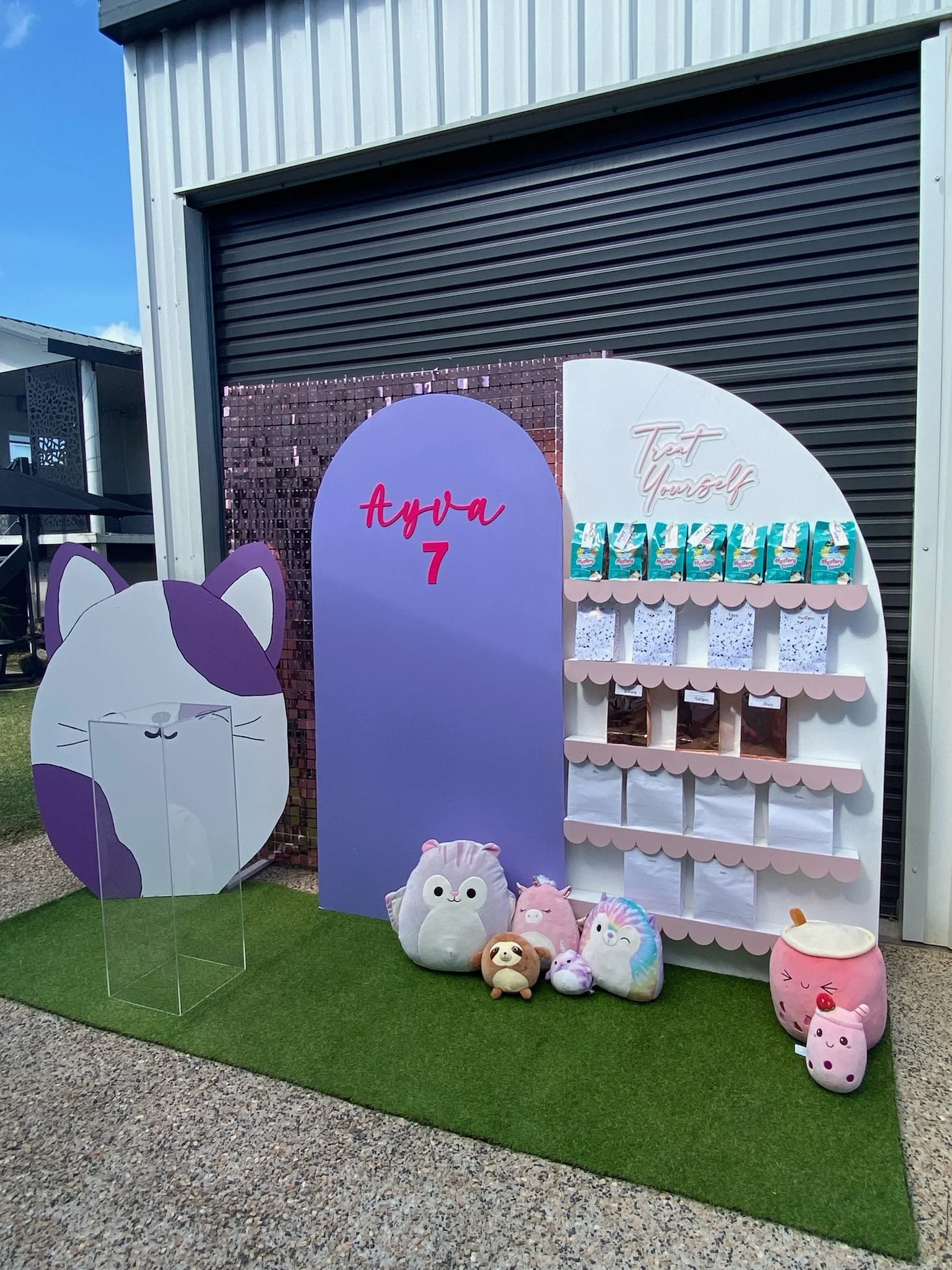 A decorated children's birthday party setup with plush toys, a purple sign with 'Ayaa 7' on it, a white shelving unit with snack bags, and a large purple and white cat figure on a patch of artificial grass outside a building.