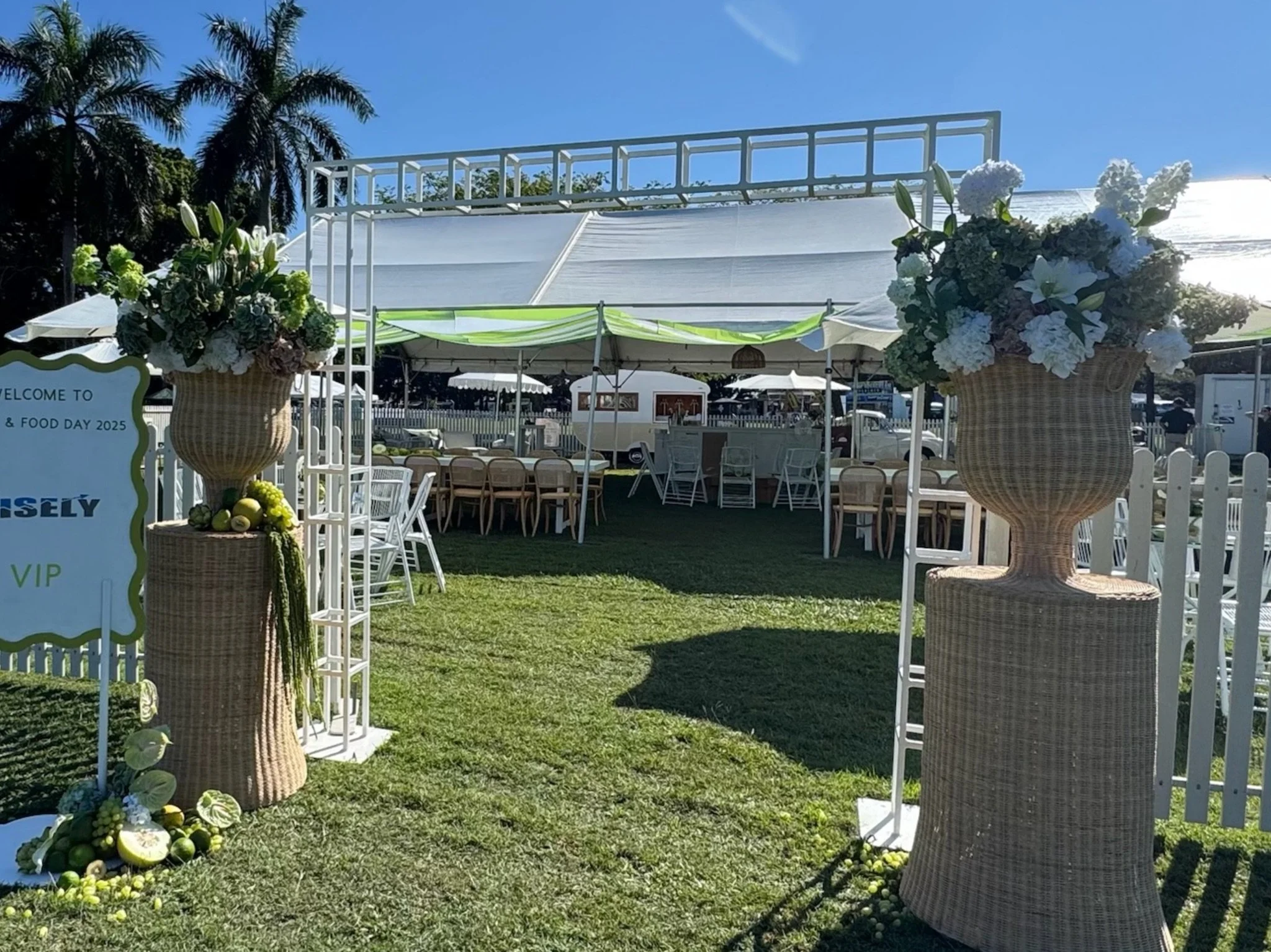 Decorated outdoor event space with large floral arrangements in woven vases, white fencing, chairs, tables, and a canopy under blue sky with palm trees in the background.