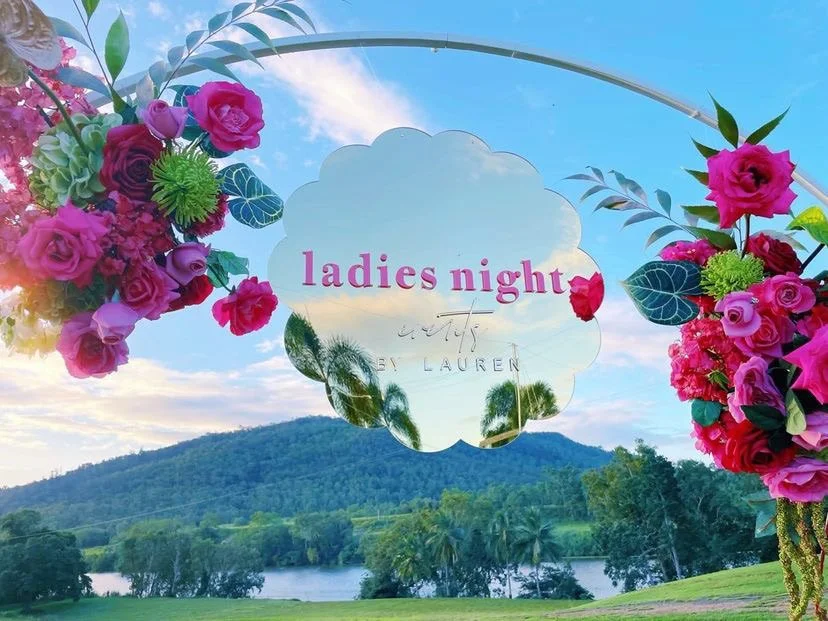 Mirror sign reading "ladies night" surrounded by pink and purple flowers with a scenic view of a lake and mountains in the background.