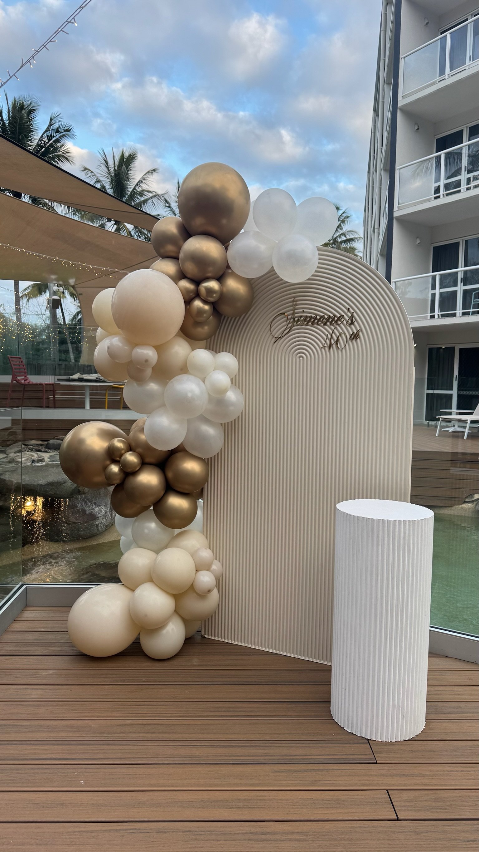 Balloon arch with white, cream, and gold balloons next to a white striped pillar and a curved backdrop with the name 'Jamaica'.