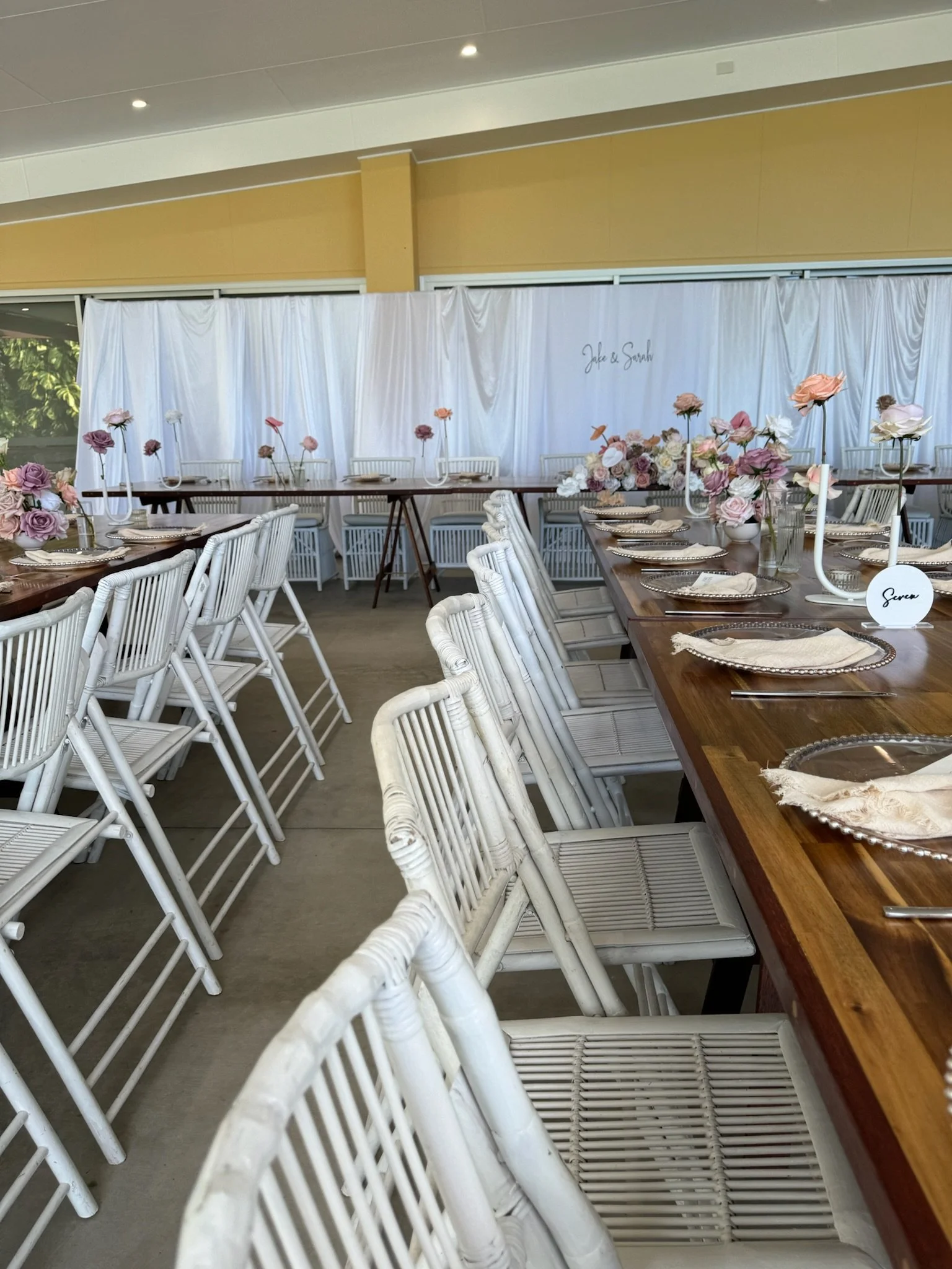 Wedding reception setup with white chairs, wooden tables, and pink floral centerpieces in vases, with a white draped backdrop displaying names 'Jade & Sarah'.