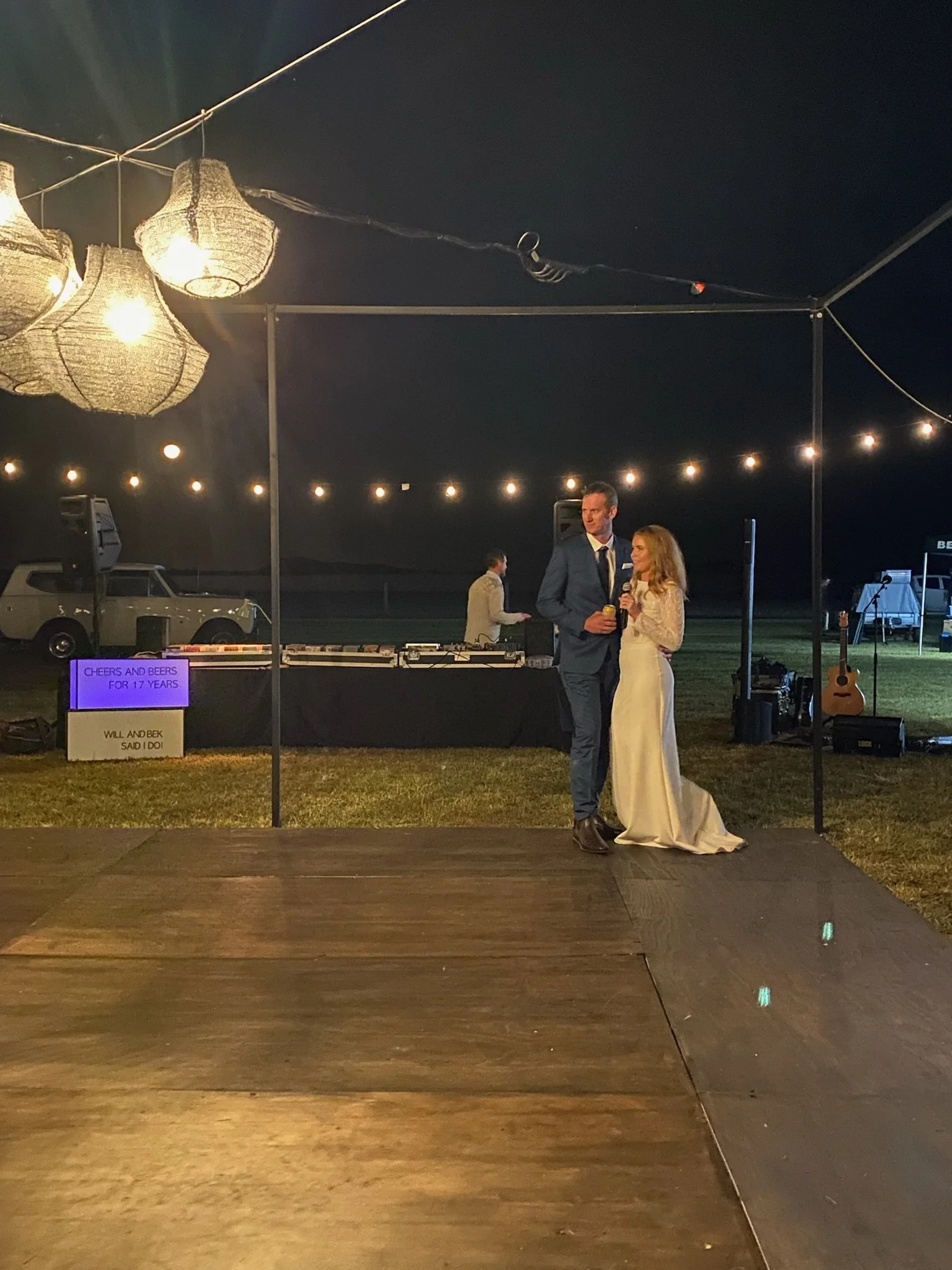 A couple dressed in formal attire, standing on a small stage at an outdoor nighttime wedding reception, with a DJ booth and string lights behind them, and a guitar on a stand nearby.