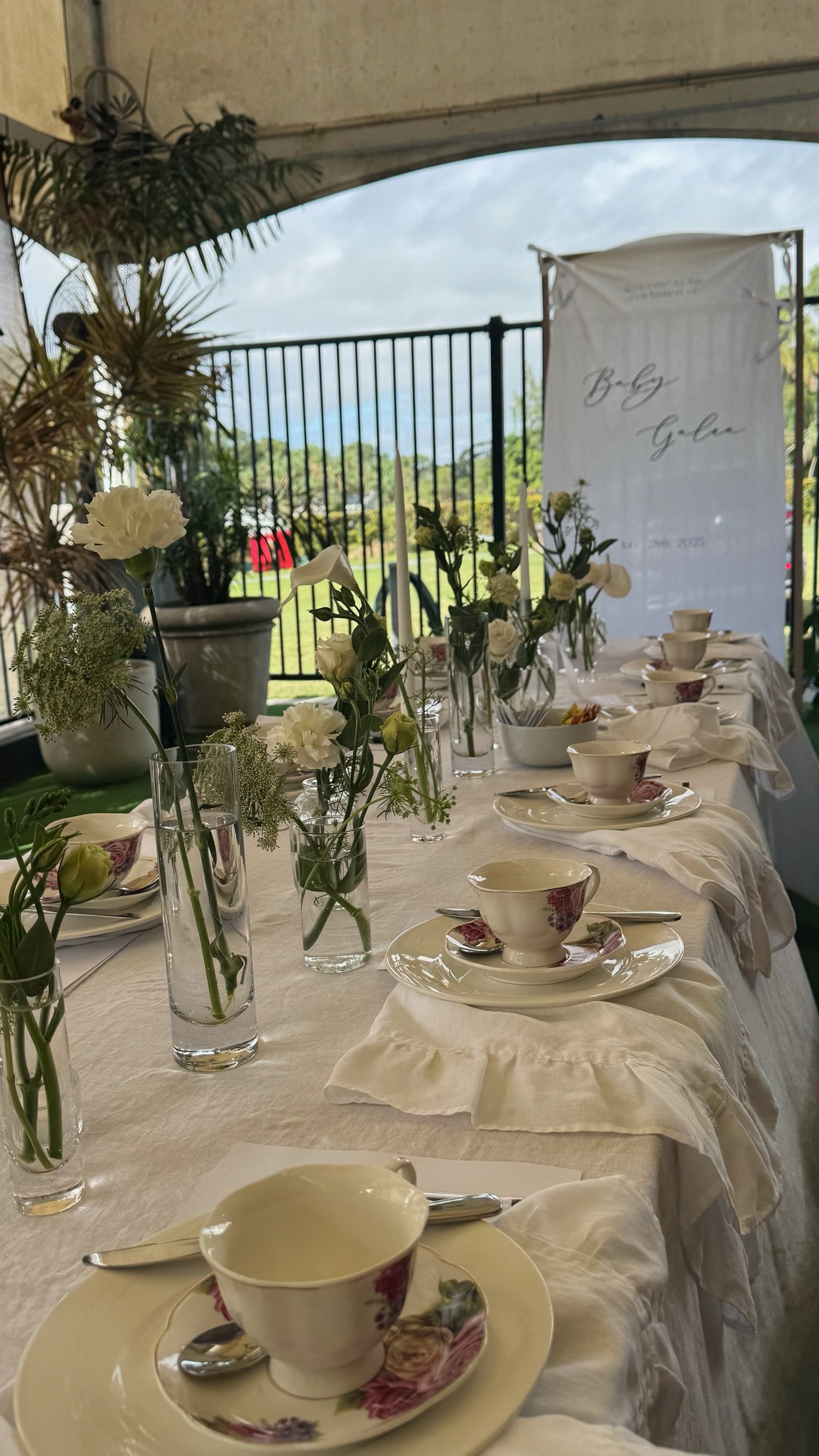 A decorated table set for a celebration with white flower arrangements in vases, tea cups on saucers, and a backdrop with the words 'Baby Gabi' and a date, likely for a baby shower or celebration.