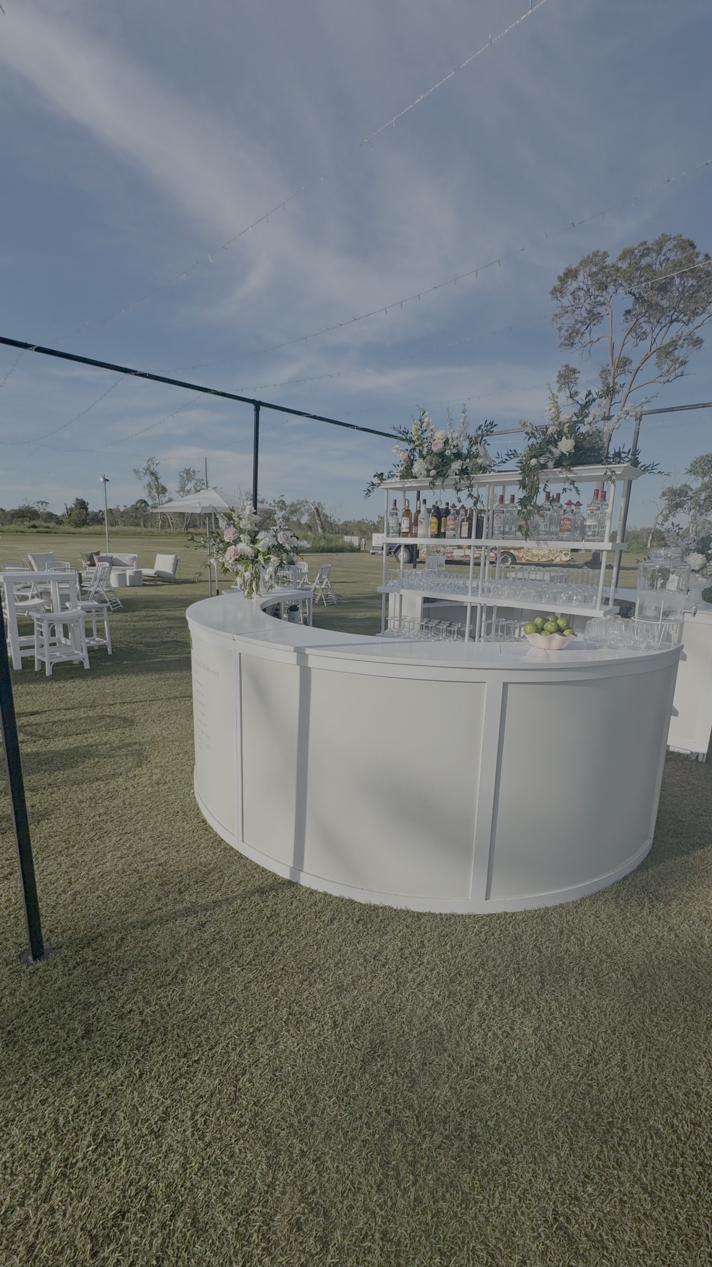 An outdoor event setup featuring a white curved bar with floral arrangements and a backdrop of open grassy field and trees under a partly cloudy sky.