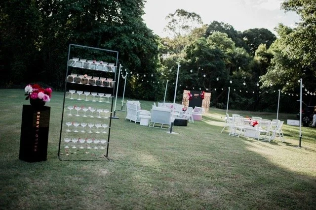 Outdoor wedding reception setup with white chairs, tables, and string lights in a grassy area surrounded by trees.