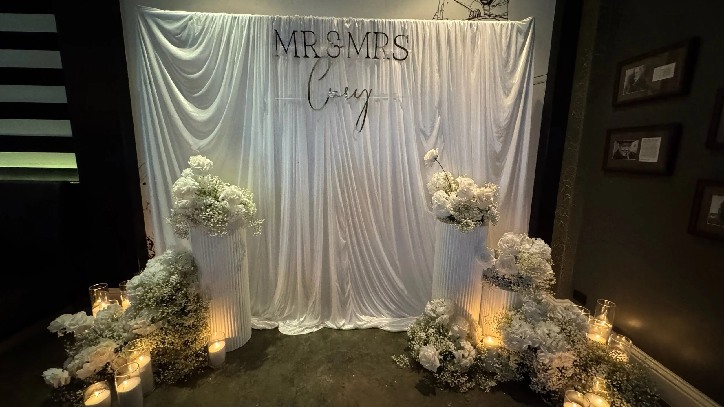 White draped backdrop with 'MR & MRS' and 'Love' signs, surrounded by white floral arrangements and candles in glass holders at a wedding celebration.