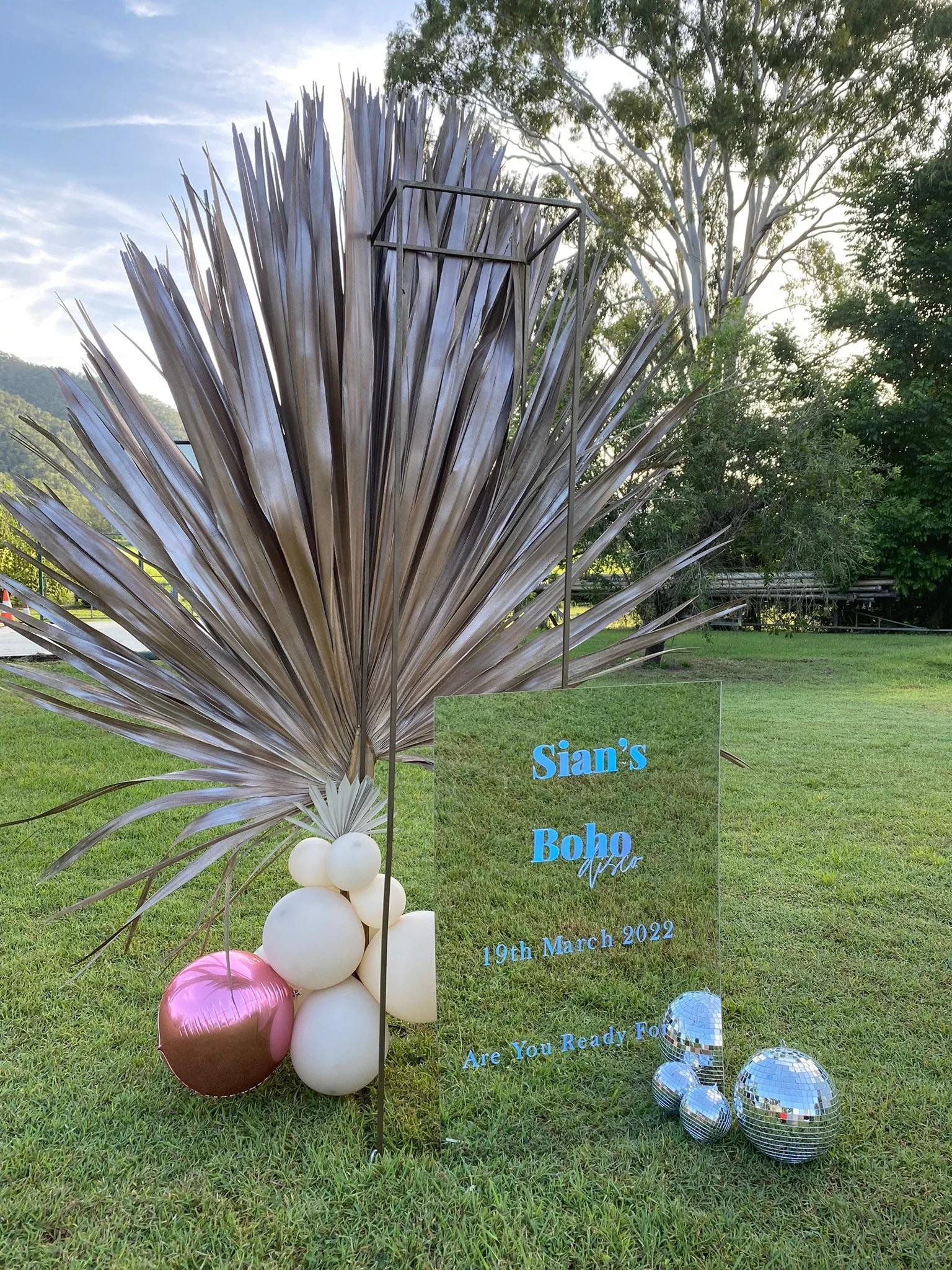 Decorative outdoor setup with metallic palm leaf, balloons, and a glass sign that reads 'Sian's Boho' with date '19th March 2022' and the phrase 'Are You Ready For'.