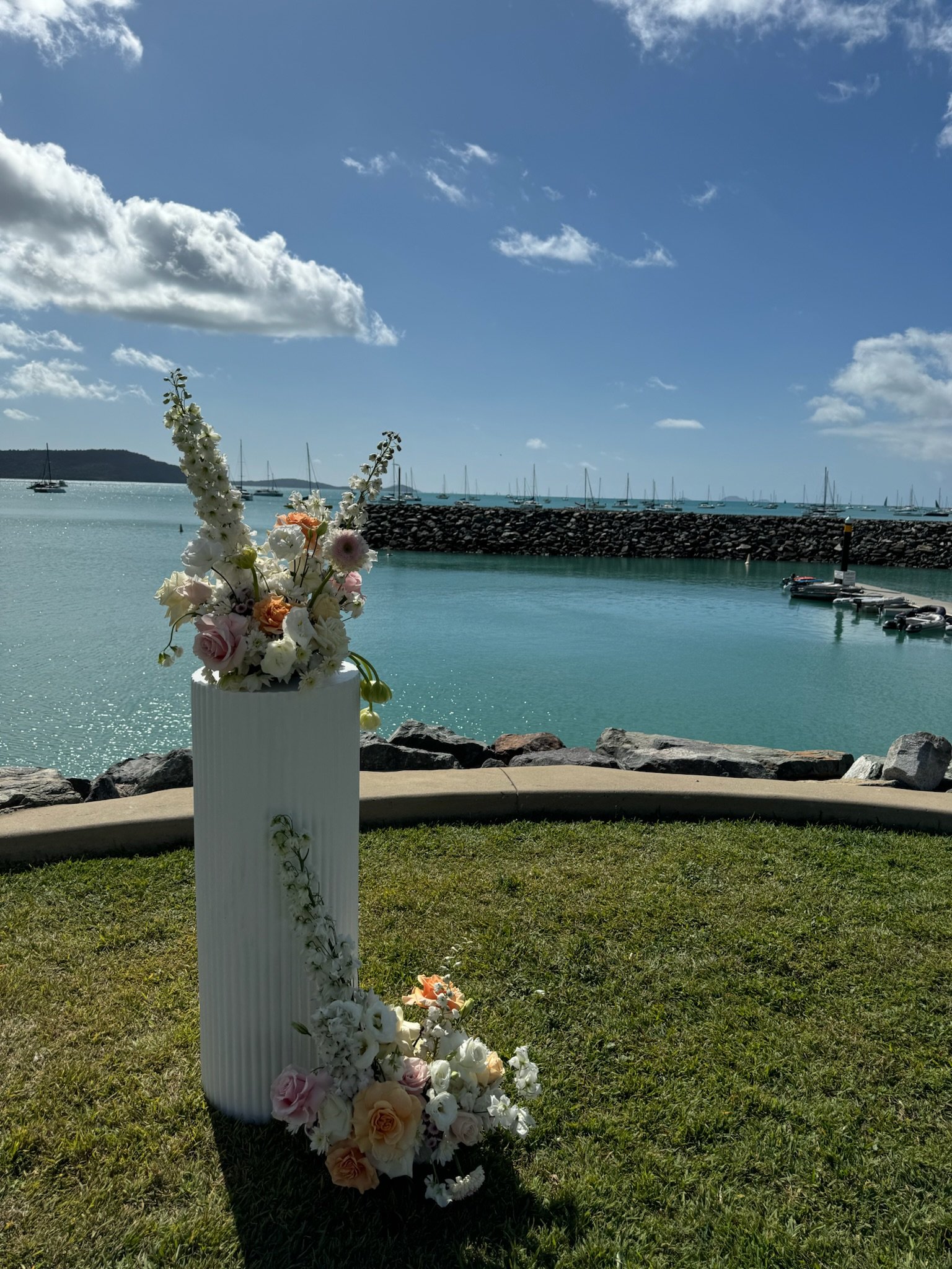 A vase with white, pink, and orange flowers on a grassy area near a harbor with sailboats, rocky breakwater, and blue sky with clouds