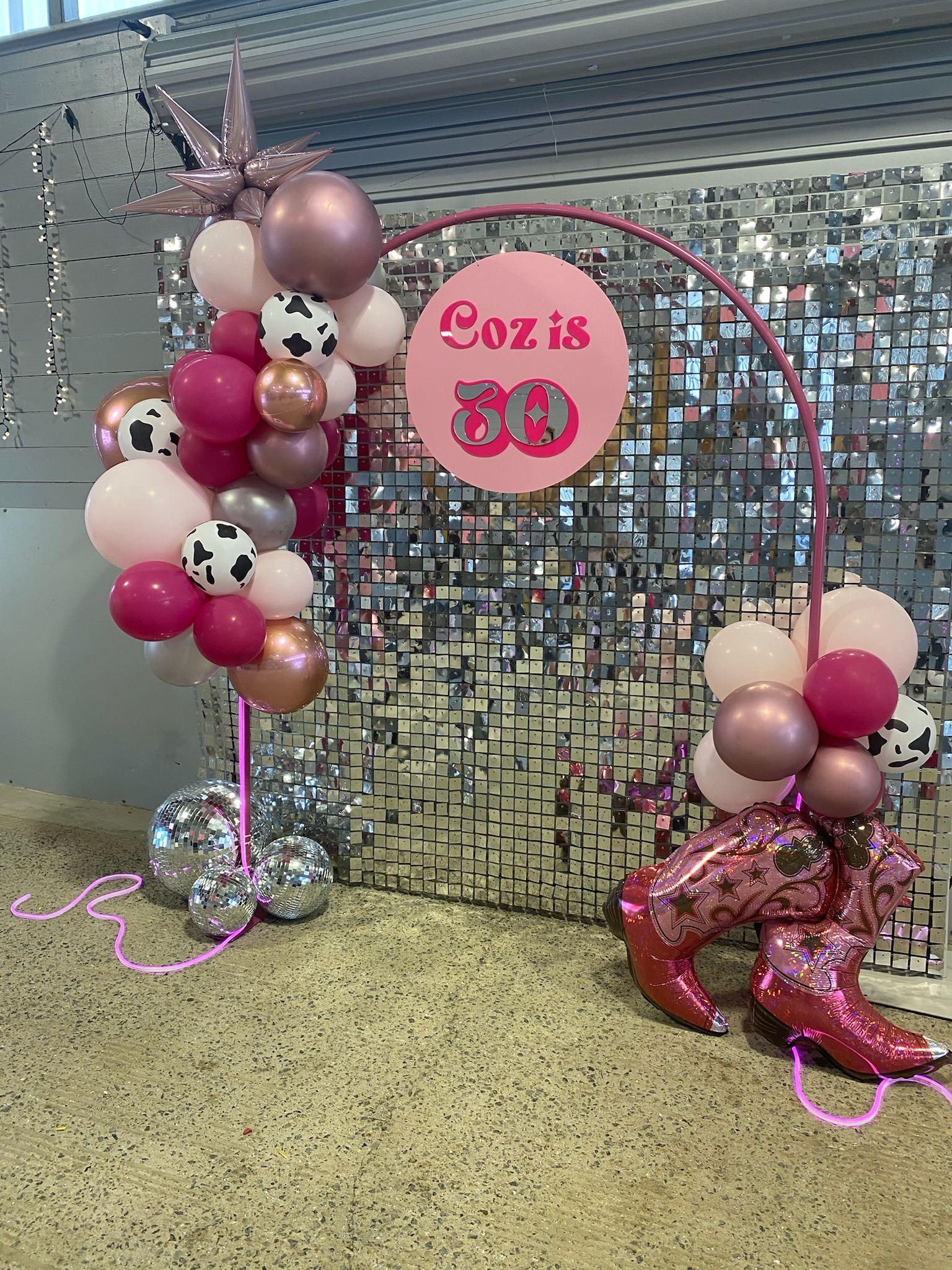 Decorative balloon arch with pink, white, gold, and cow print balloons, a pink sign reading "Coz is 30," and pink glittery cowboy boots balloons, set against a metallic sequin backdrop.