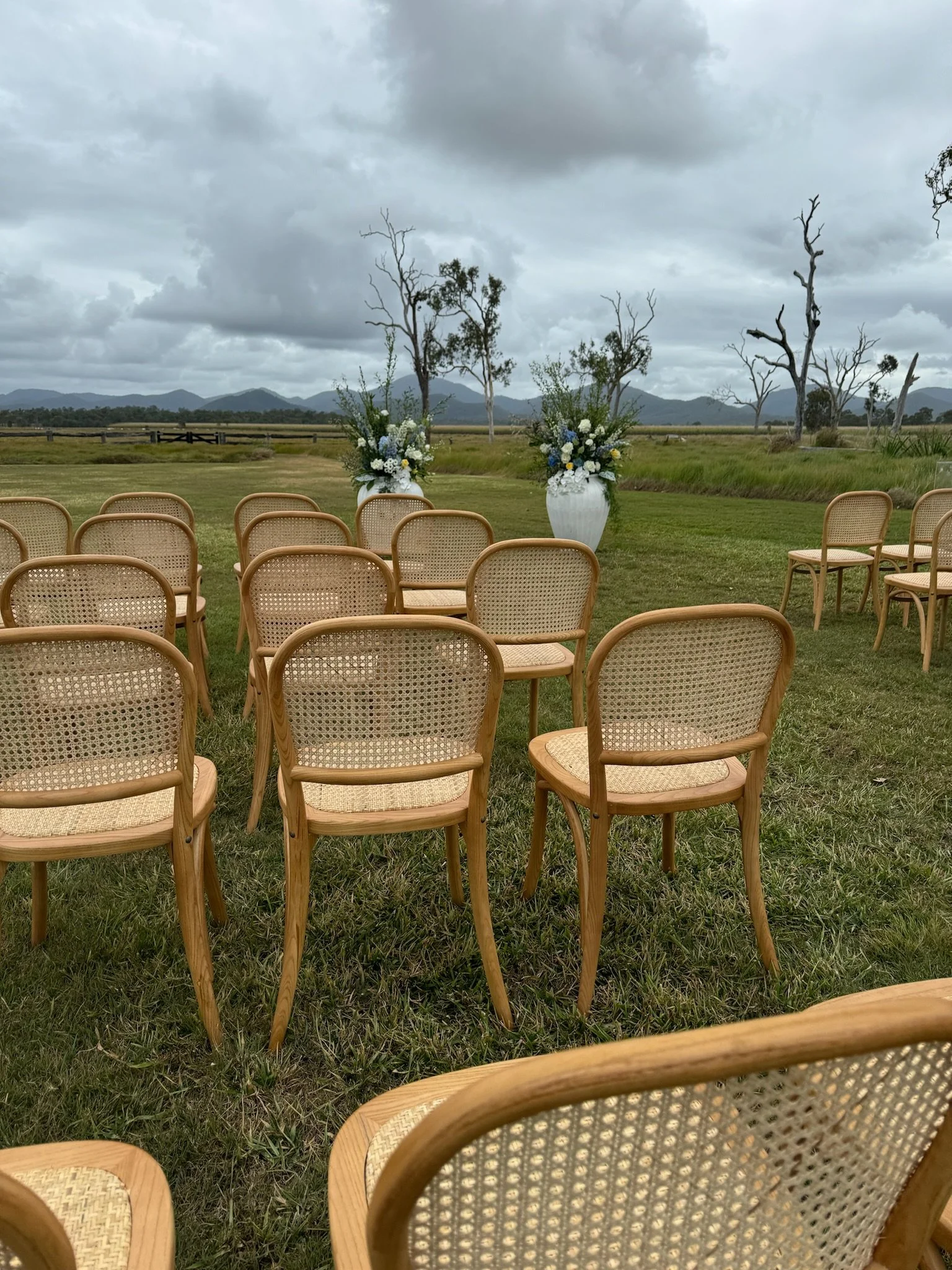 Outdoor wedding setup with wooden chairs arranged on grass, large vases with flowers, and a rural landscape background with mountains and leafless trees under cloudy skies.