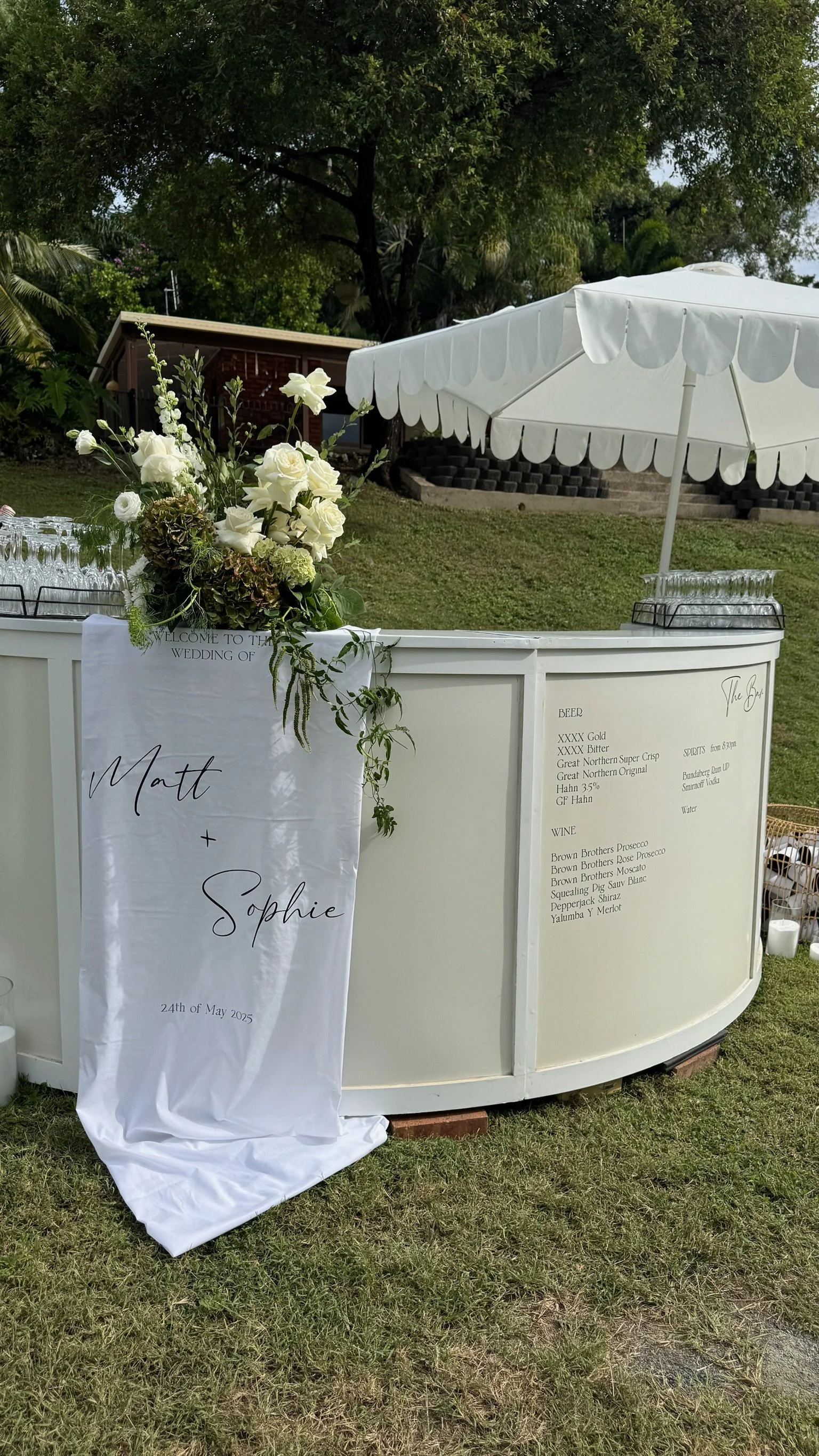 Wedding reception drink station with a white curved bar, floral arrangements, and a white umbrella in an outdoor garden setting.