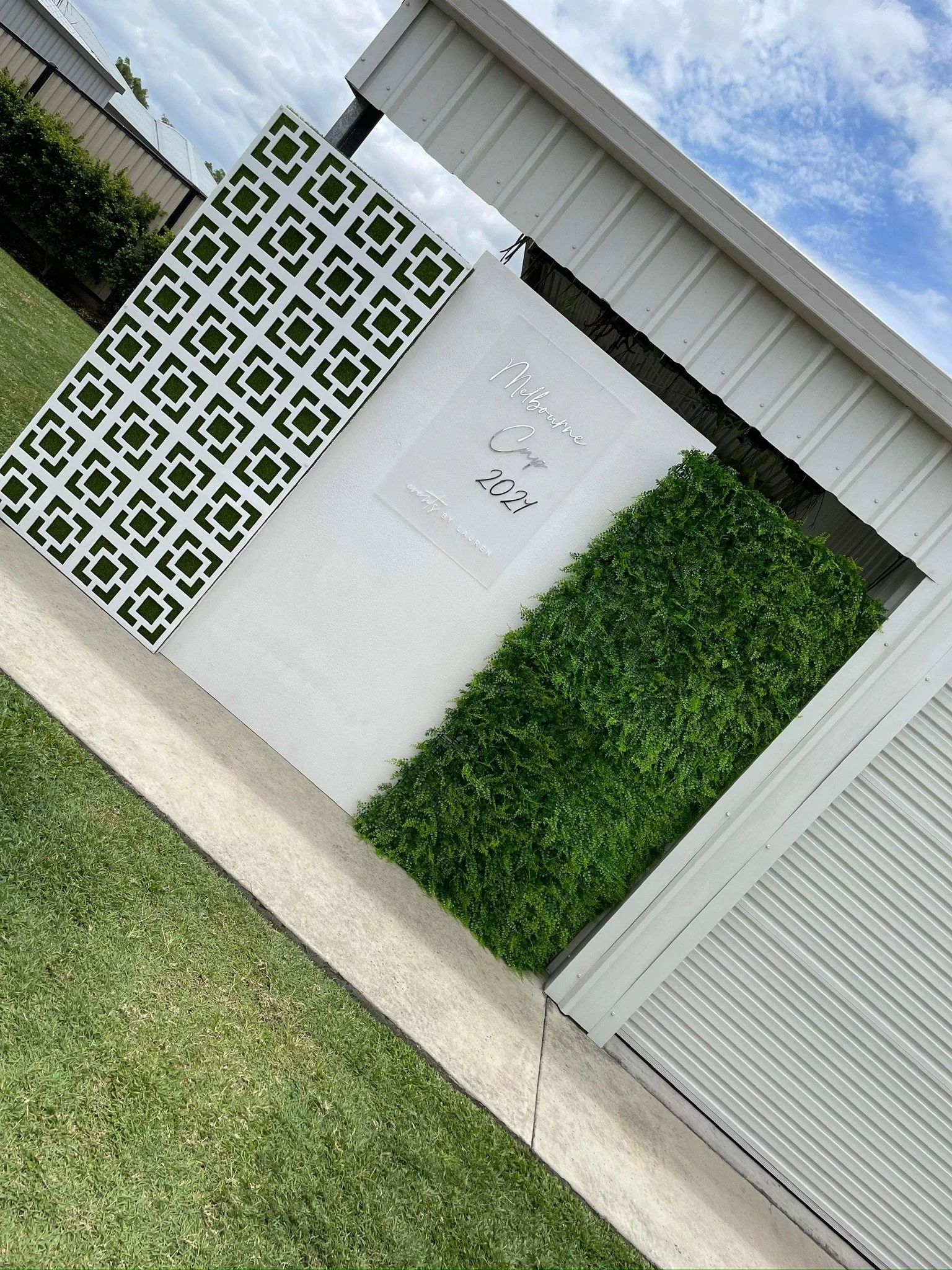 A decorated outdoor space with a white lattice panel, a white sign with handwritten text, a green hedge wall, and a concrete walkway, under a cloudy sky.